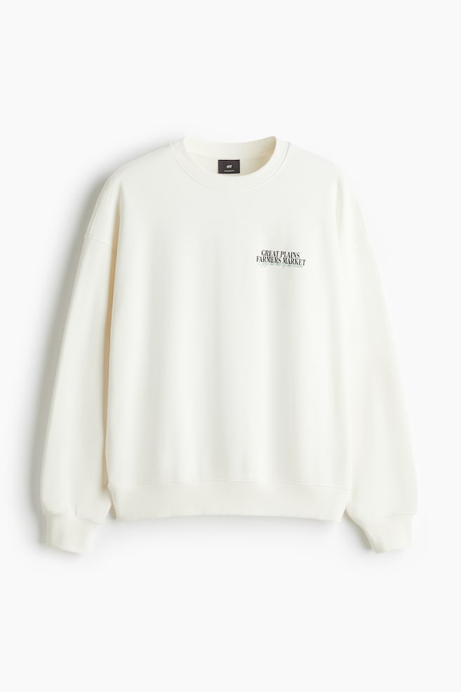 Ein cremefarbenes Sweatshirt aus mittelschwerem Baumwollstoff wird von vorne präsentiert und zeigt seine übergroße, lockere Passform mit überschnittenen Schultern, einem runden, gerippten Halsausschnitt, gerippten Bündchen und einem gerippten Saum. Ein schwarzes 'GREAT PLAINS FARMERS MARKET'-Druckmotiv ist auf der oberen linken Brust sichtbar. - 1