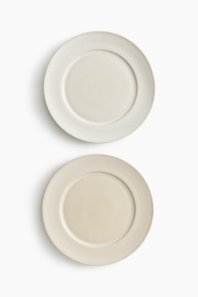 Two round ceramic plates are shown from a front overhead view, arranged vertically. Each plate features a speckled, matte glaze in light off-white and beige tones, with a wide rim and a recessed centre. - 1