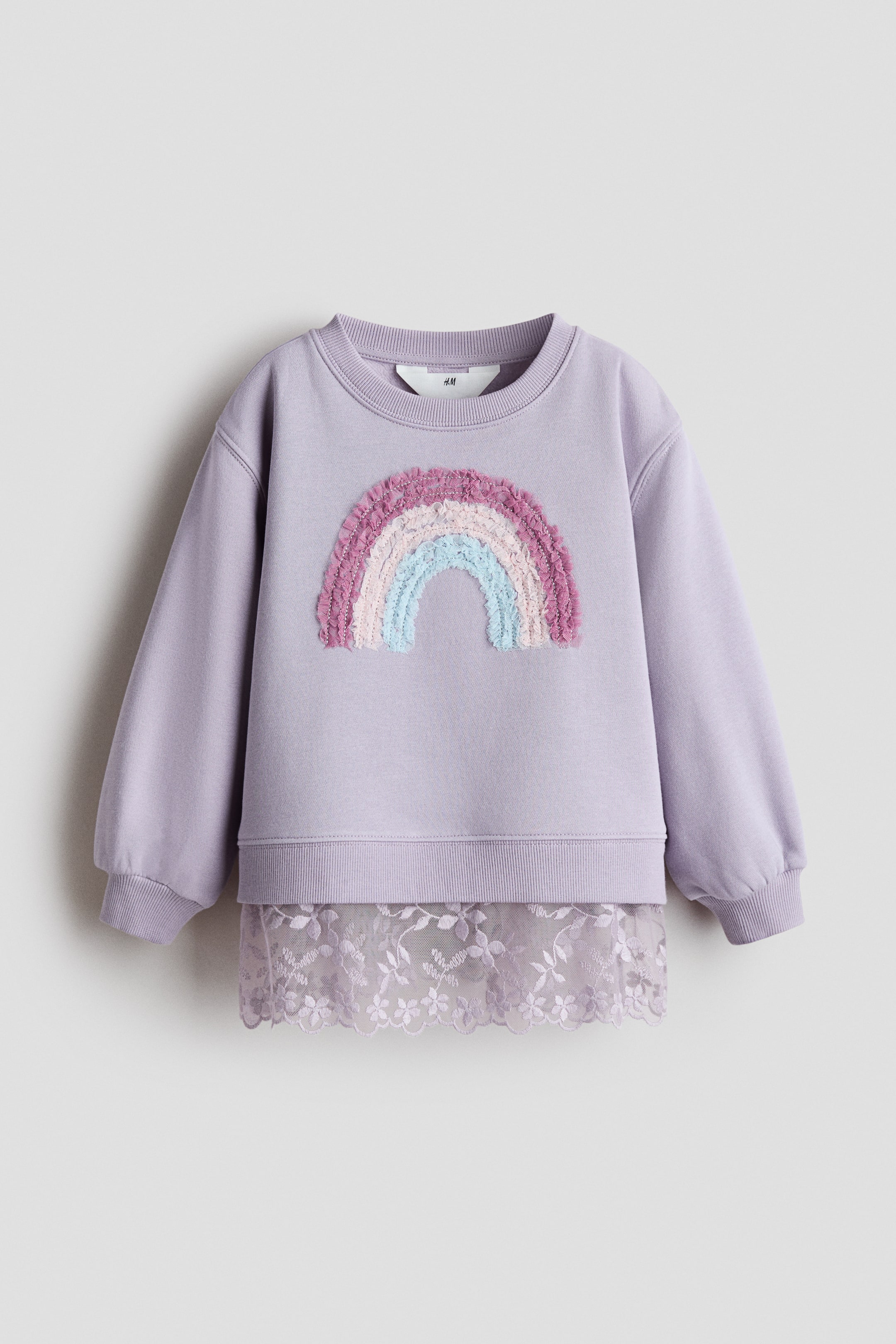 View larger image: A light purple jumper is showcased from the front, highlighting a textured rainbow appliqué across the chest with bands of fuchsia, pink, light blue, and light green. The jumper features a ribbed crew neck, ribbed cuffs, and a ribbed hem, with a light purple floral lace hem peeking from underneath.