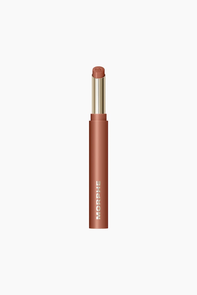 En matt rødlig-brun leppestifthylse står oppreist, og viser frem en forlenget lys brun leppestiftkule. Kulen stiger fra et gulltonet indre rør, og et 'M'-logo er preget på spissen. Merkenavnet 'MORPHE' er trykt vertikalt på forsiden av det ytre røret. - 1