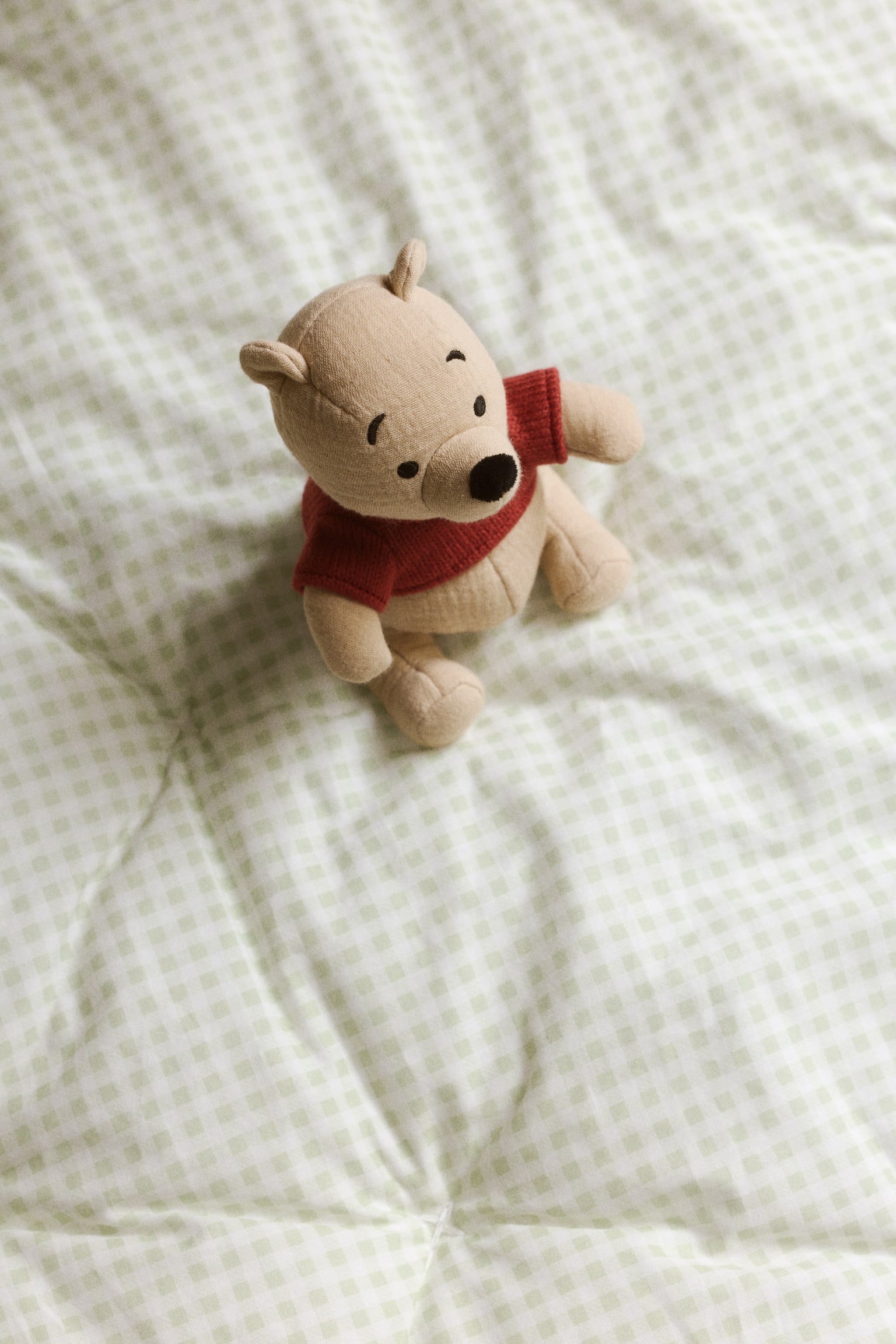 A beige bear-shaped rattle is sitting, facing front, featuring embroidered black eyes, nose, and mouth, and wearing a red knitted top. - 1