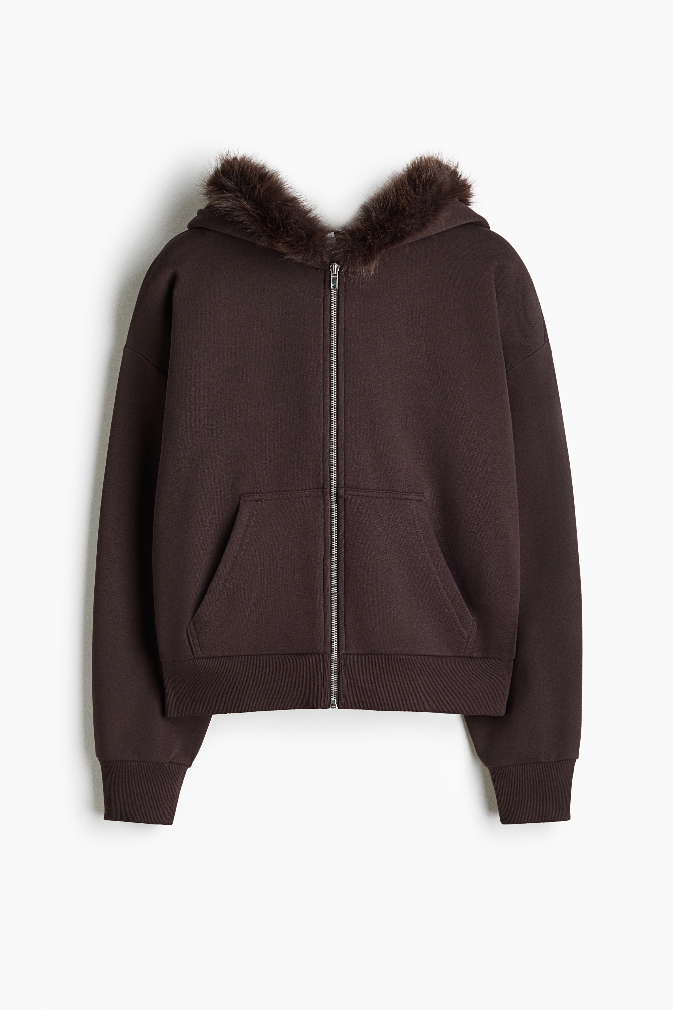 A dark brown zip-up fleece hoodie featuring a hood with a fluffy faux-fur trim is being worn by the model, who is adjusting the top of the hood with one hand. The loose-fit hoodie has dropped shoulders, front pockets, and ribbed cuffs, and is styled over a white T-shirt and beige drawstring trousers.