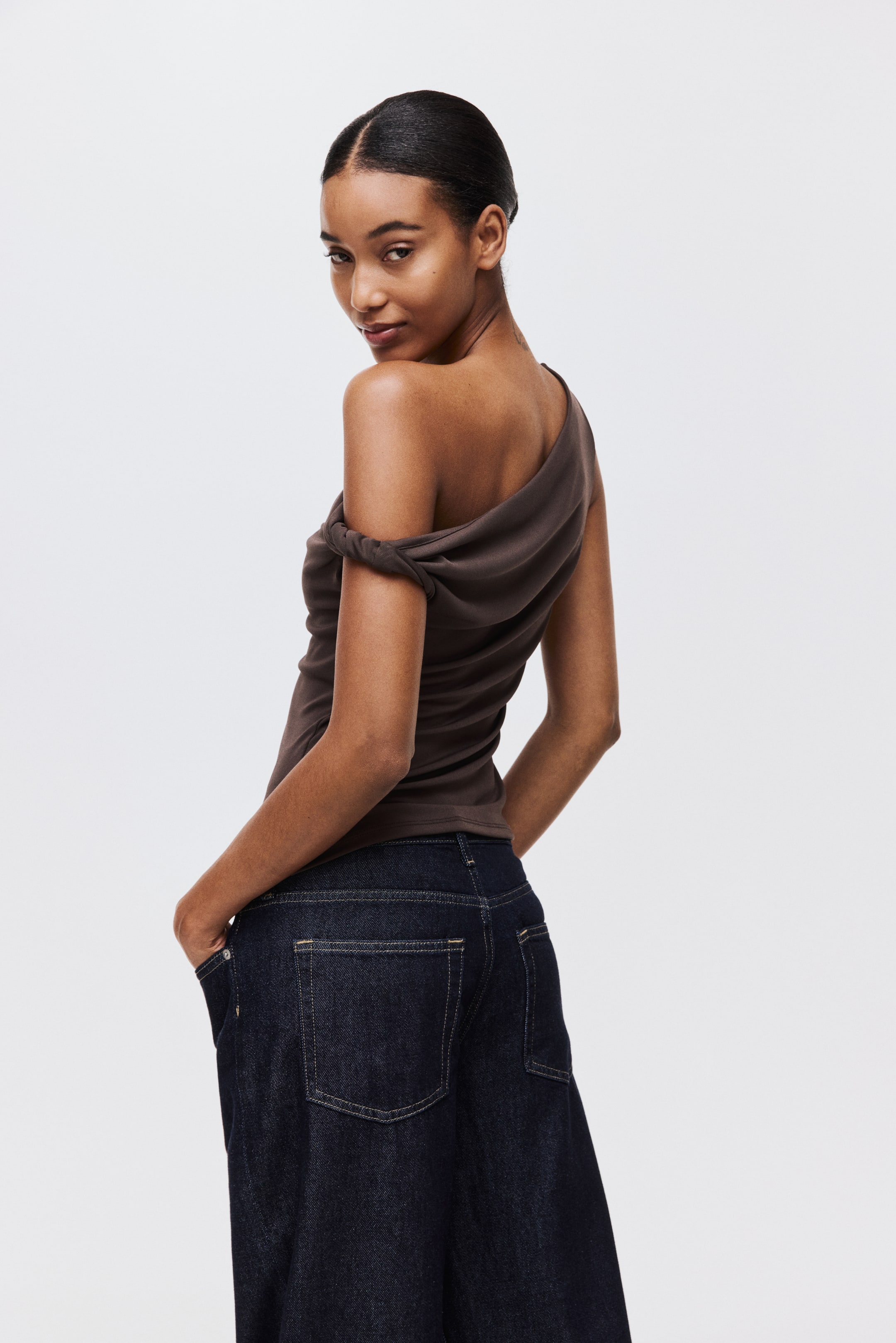 View larger image: A dark brown one-shoulder top is being displayed from a rear-side view, featuring a twisted strap on the right shoulder and an exposed left shoulder. Dark wash, wide-leg denim jeans, with visible back pockets, are being worn.