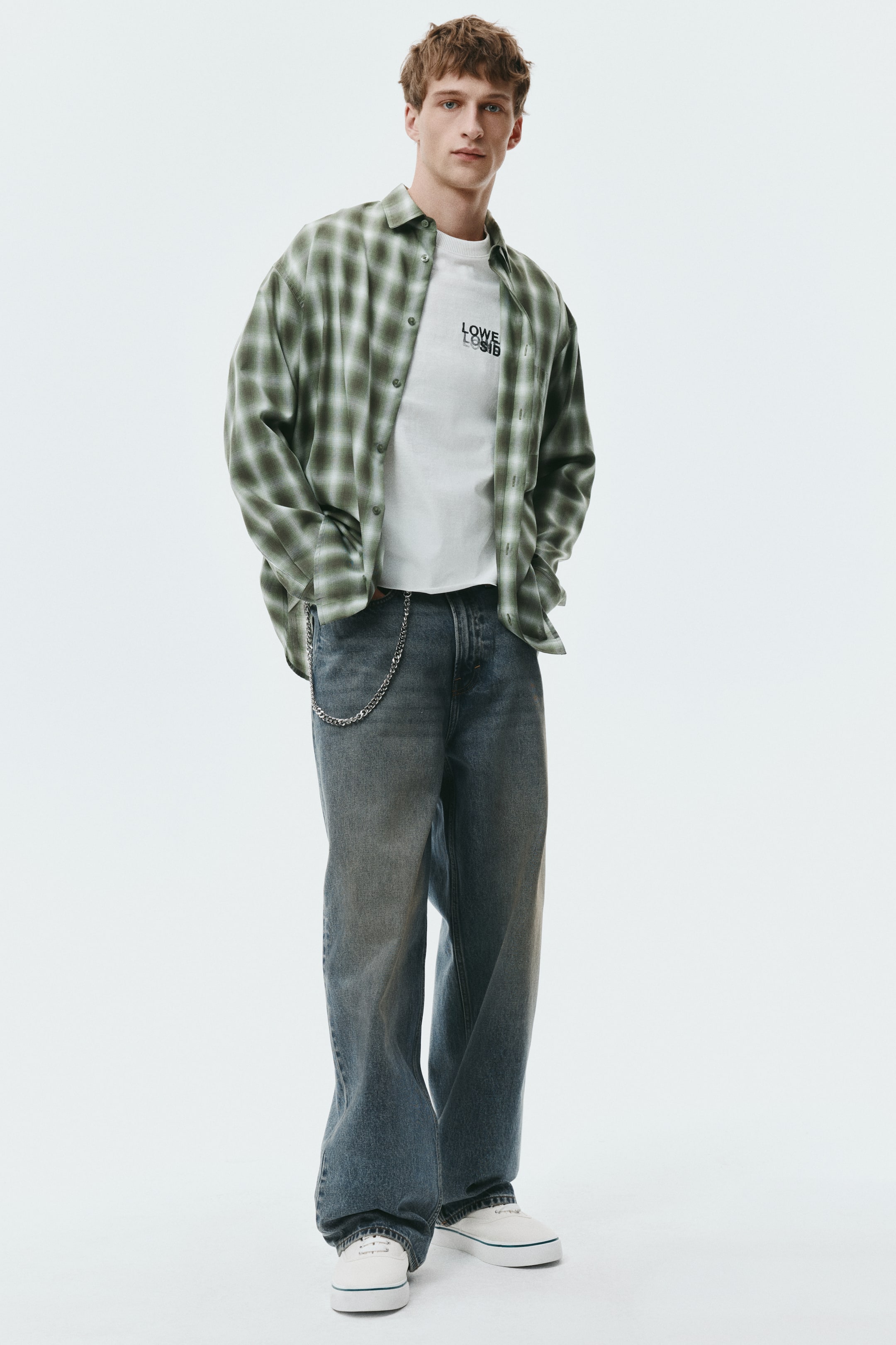 Xem ảnh lớn hơn: Quần jeans five-pocket bằng rigid denim màu xanh‑xám phai đang được người mẫu mặc. Quần có phom baggy với ống tròn và đáy quần hạ thấp (dropped crotch), vải xếp chồng ở cổ chân. Một sợi xích bạc treo trên belt loop của quần.