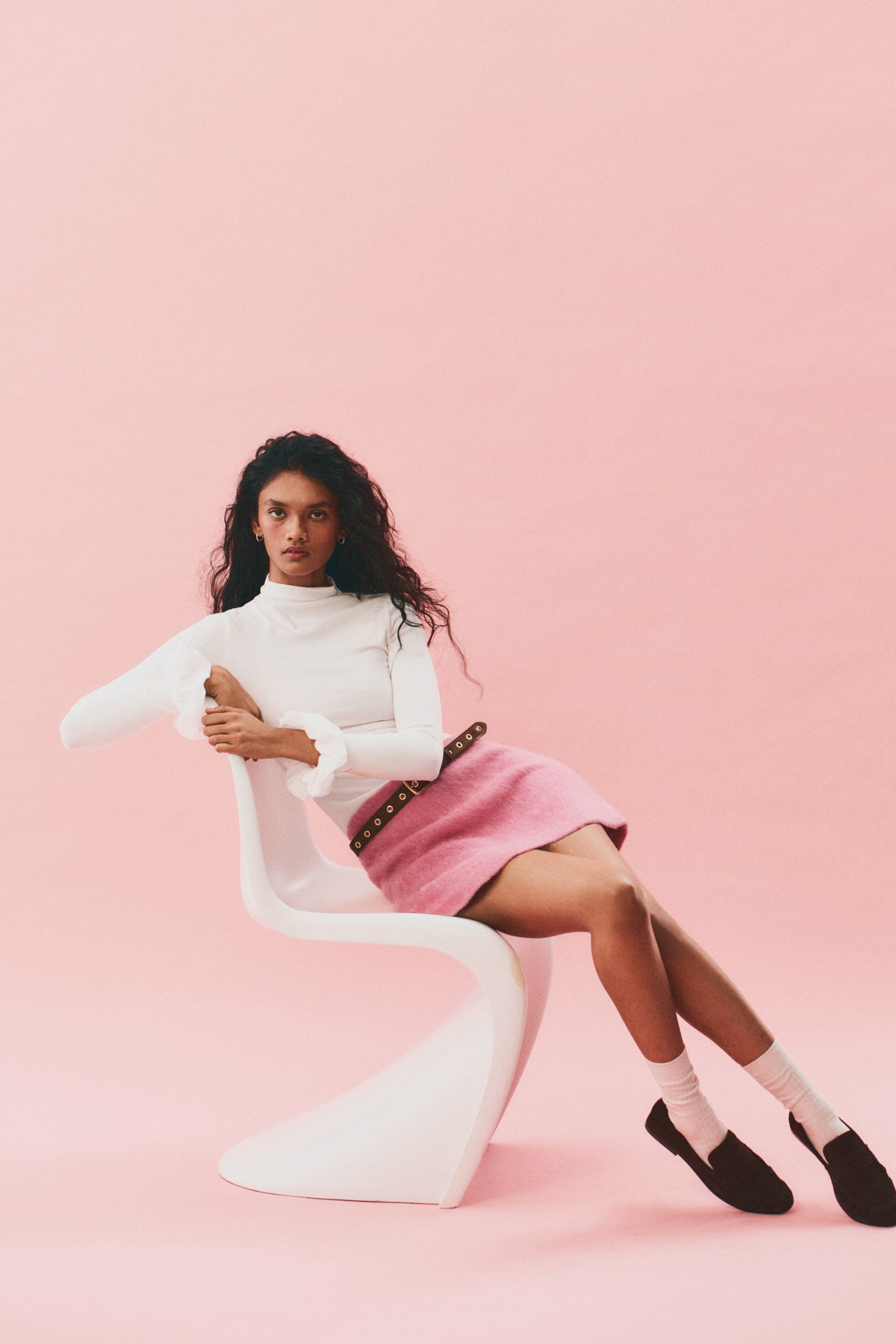 View larger image: A white long-sleeved top featuring a mock neck and ruffled cuffs is being worn, tucked into a pink mini skirt. The skirt is being accessorized with a dark belt featuring metal eyelets, and black loafers are being worn with white socks.