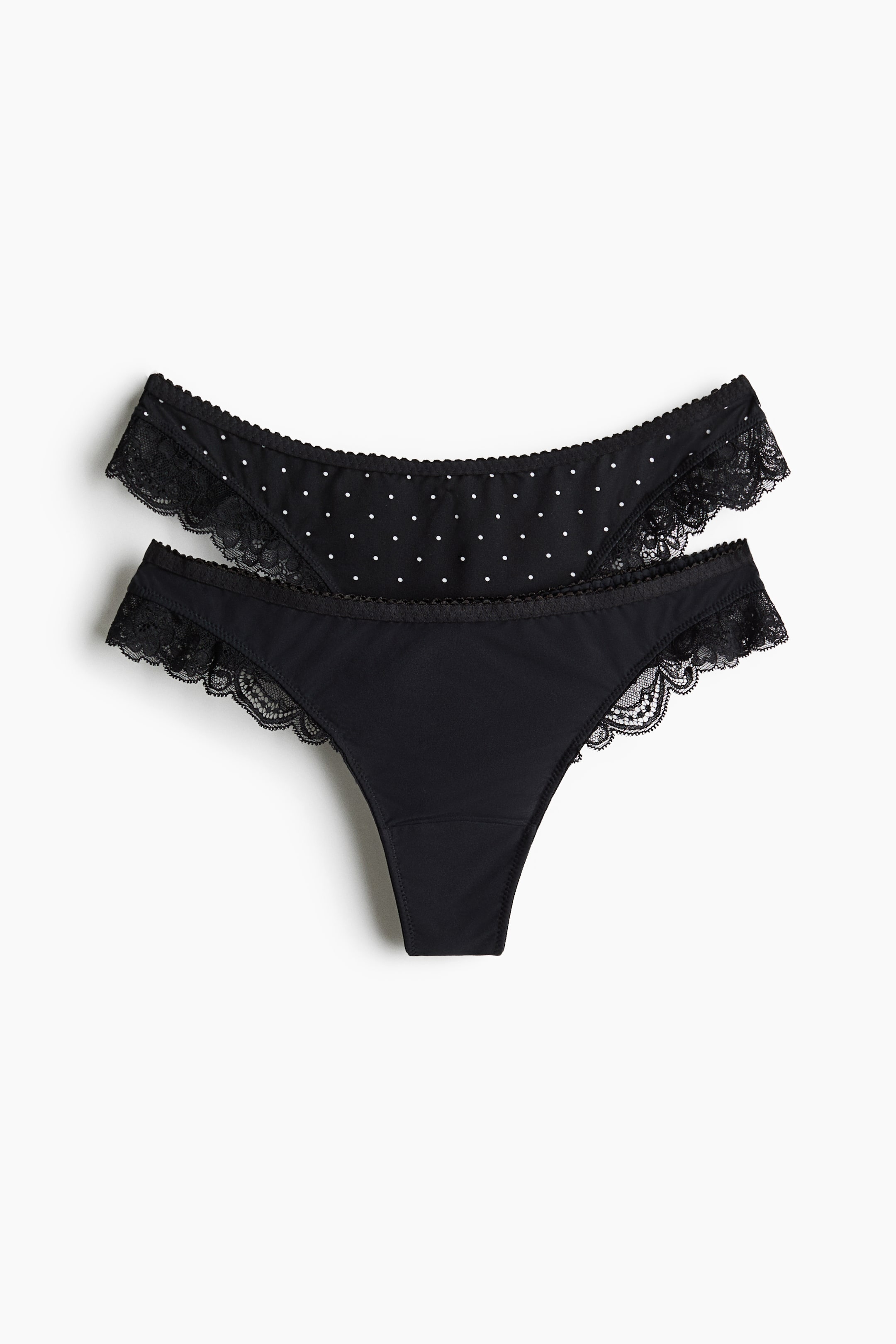 Grotere afbeelding bekijken: Een set van twee zwarte slips is van voren afgebeeld, met één effen zwarte slip en één slip met een wit polkadotpatroon. Beide designs zijn voorzien van een zwart geschulpte kanten afwerking langs de beenopeningen en een smalle, elastische tailleband.