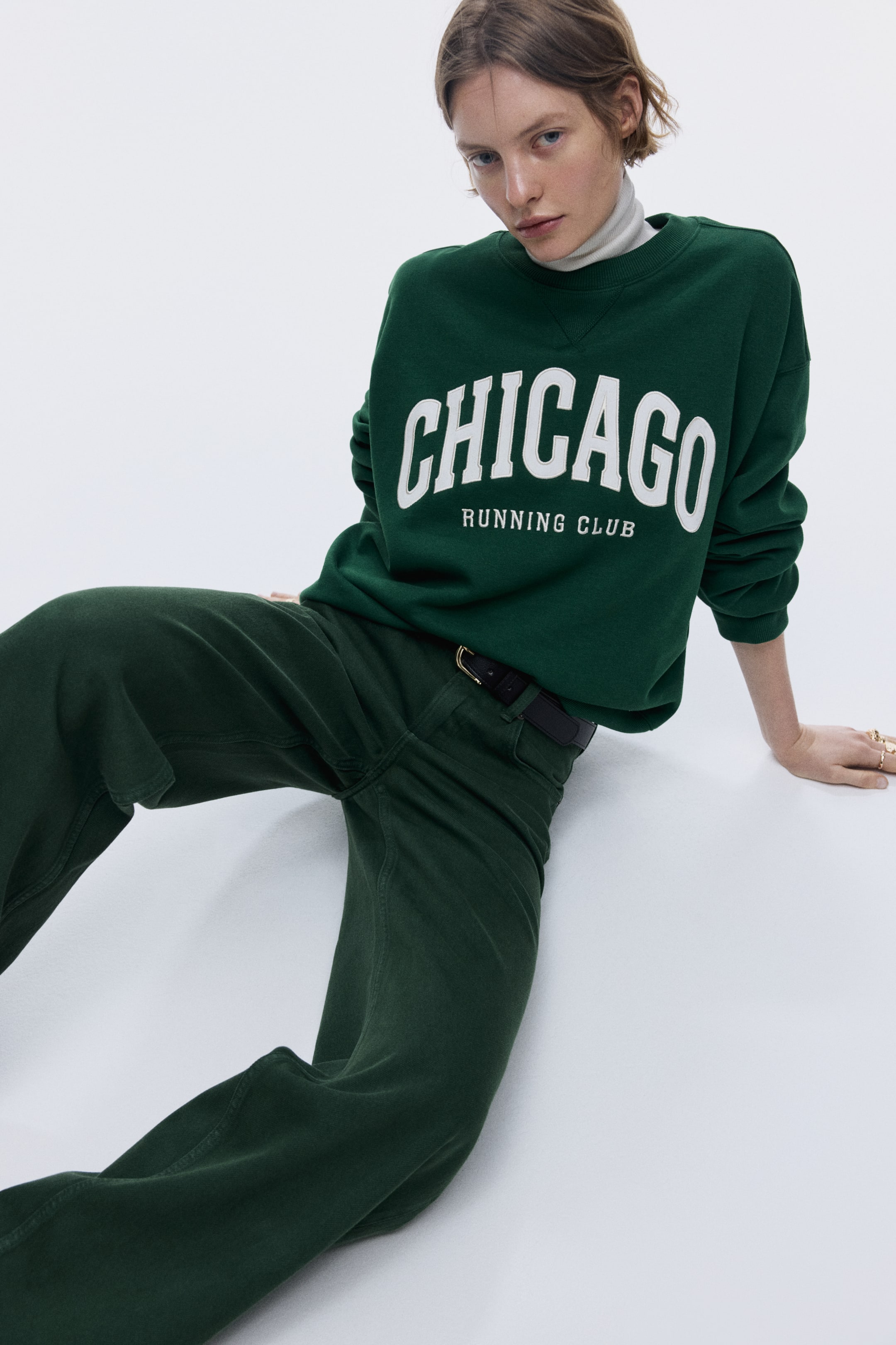 View larger image: A dark green sweatshirt is displaying white 'CHICAGO RUNNING CLUB' text on its front, featuring a ribbed crew neck and a white turtleneck layered underneath. It is being worn with dark green, wide-leg trousers, positioned at a slight angle, and secured by a black belt with a gold buckle.