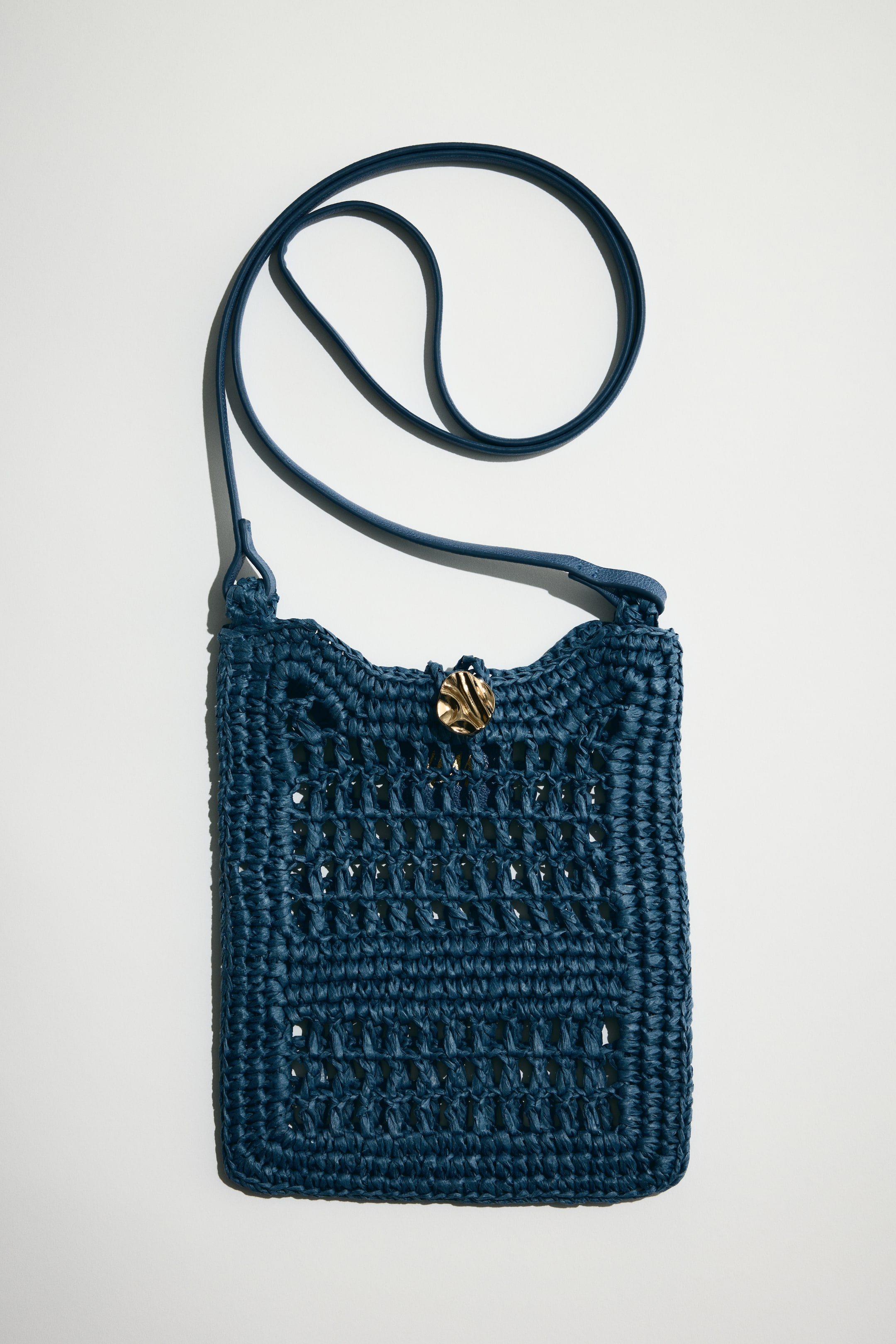 View larger image: This phone bag showcases an intricate openwork pattern crafted from dark blue paper straw, complemented by a slim shoulder strap and a metal button closure at the top.
