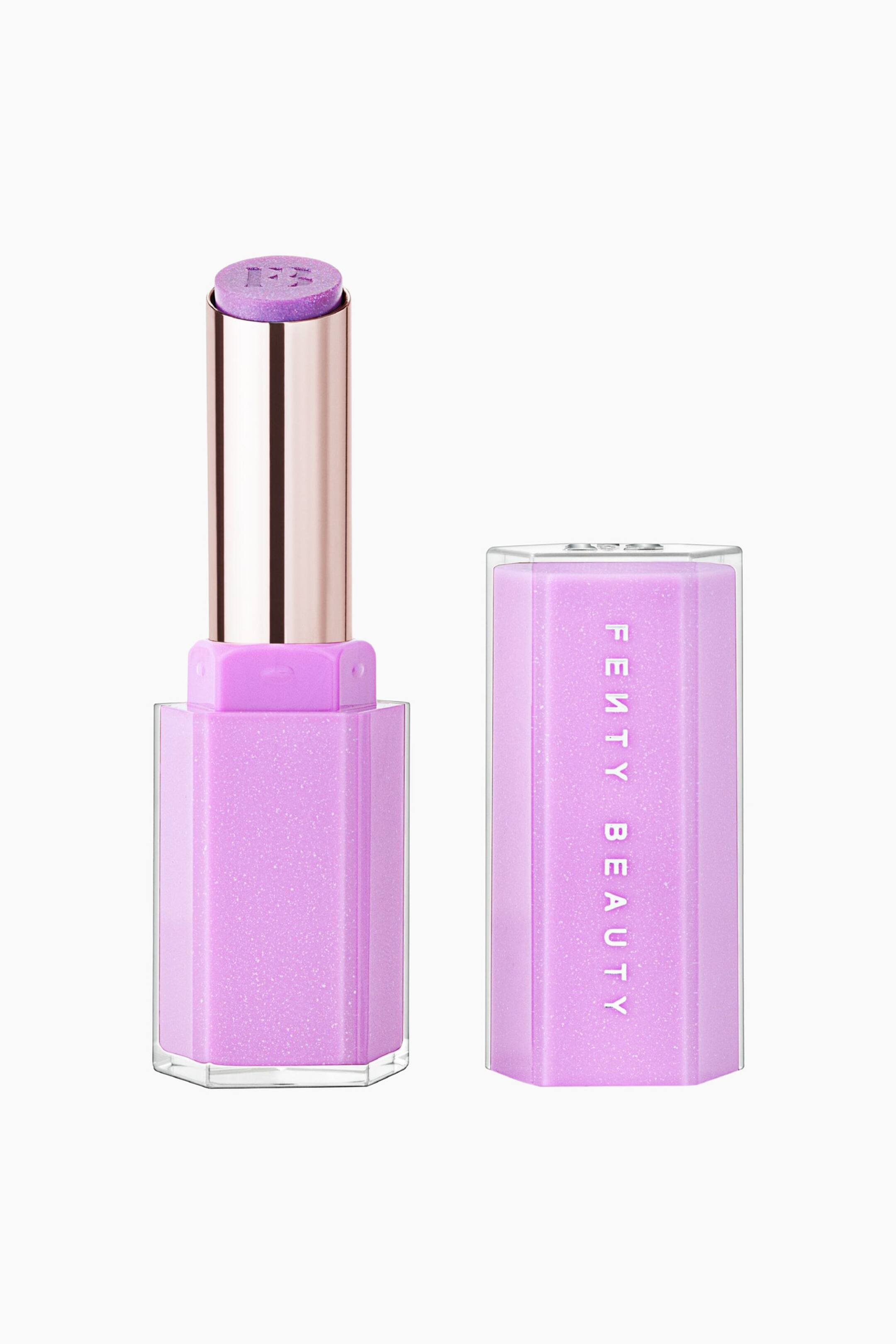 Näytä suurempi kuva: Gloss Bomb Stix High-shine Shimmer Gloss Stick -huulikiilto on pystyasennossa irrotetun korkkinsa vieressä, ja siinä näkyy kuusikulmainen kirkas kotelo, jonka sisällä on hohtava vaaleanvioletti ydin. Avoin kotelo paljastaa metallinhohtoisen ruusukultaisen sylinterin, jossa on violetti shimmer-huulikiiltotikku ja siihen kohokuvioitu "FB"-logo, kun taas yhteensopivassa korkissa näkyy pystysuuntainen valkoinen "FENTY BEAUTY"-brändäys.