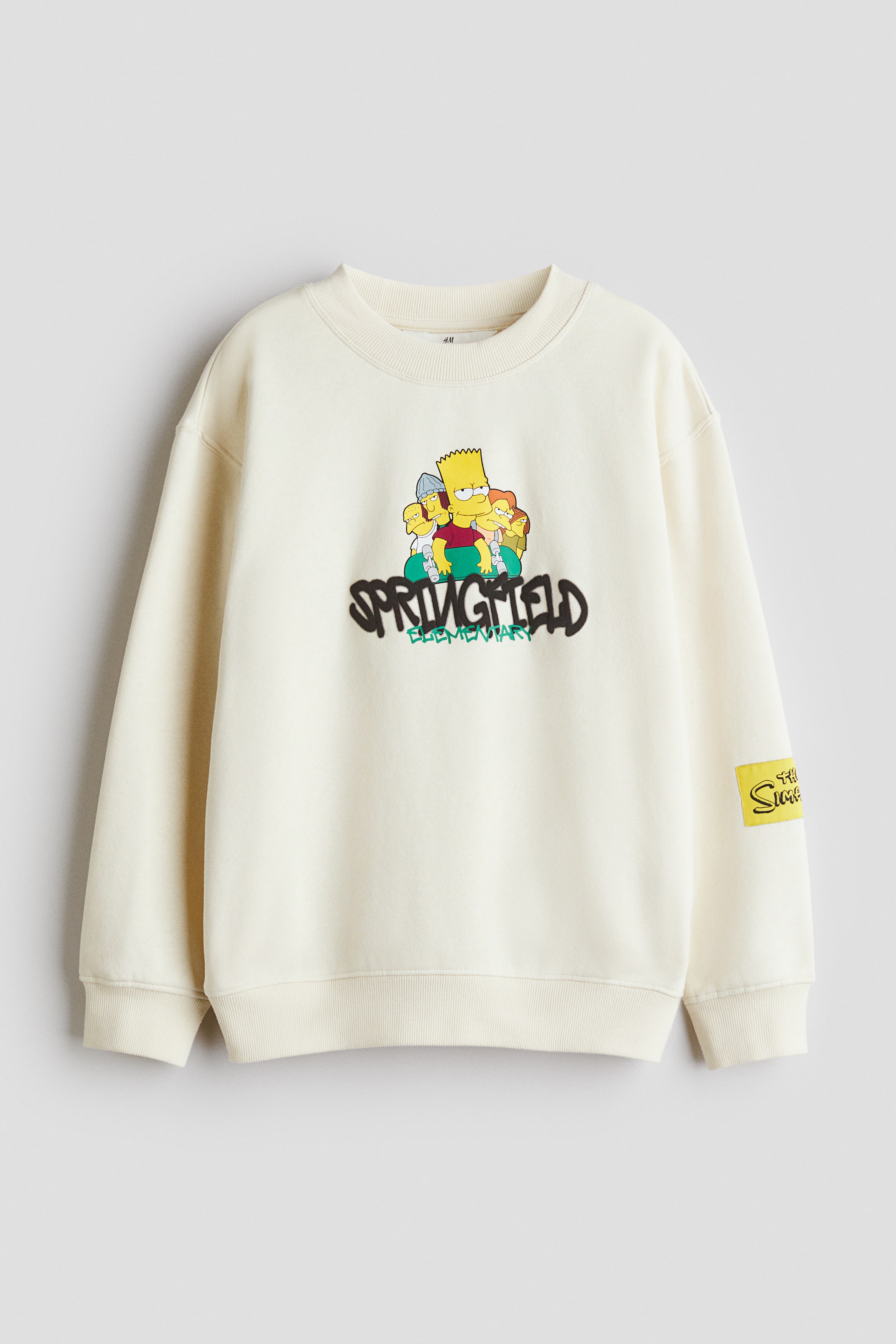 Büyük resmi görüntüle: Krem rengi bir sweatshirt, ribana bisiklet yaka, ribana manşetler ve ribana etek ucu ile sergileniyor. Ön kısmı, "Springfield Elementary" yazısının üzerinde Bart Simpson ve diğer karakterlerin yer aldığı renkli bir grafikle süslenmiş. Sol kolda "The Simpsons" logosunu taşıyan sarı dikdörtgen bir yama görünüyor.