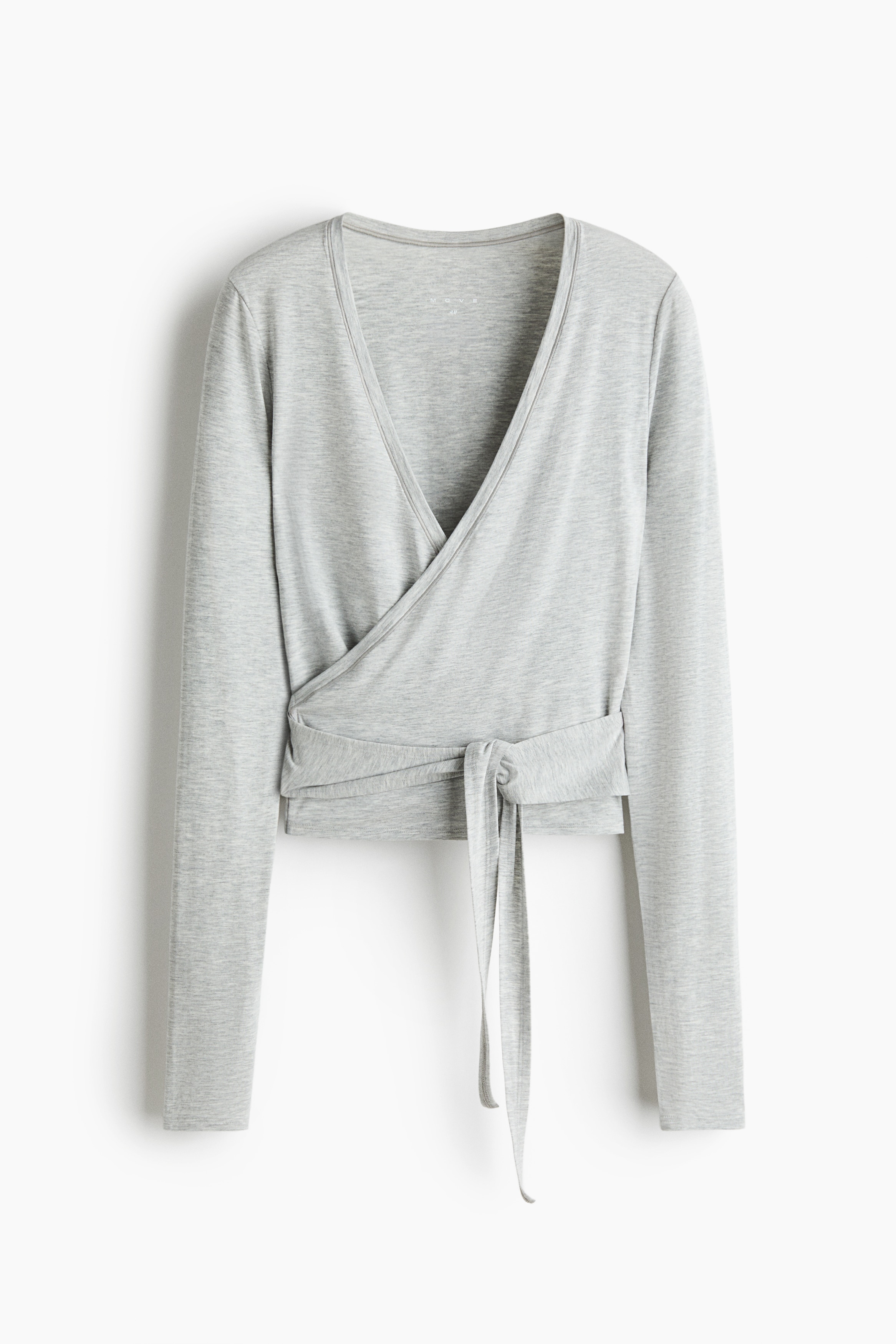 A light grey, long-sleeved wrap top in a soft jersey fabric is being worn by the model, featuring a deep V-neckline and ties wrapped around the waist. The slim-fit top is layered over a white tank top and styled with cream-coloured athletic shorts and grey ribbed leg warmers.