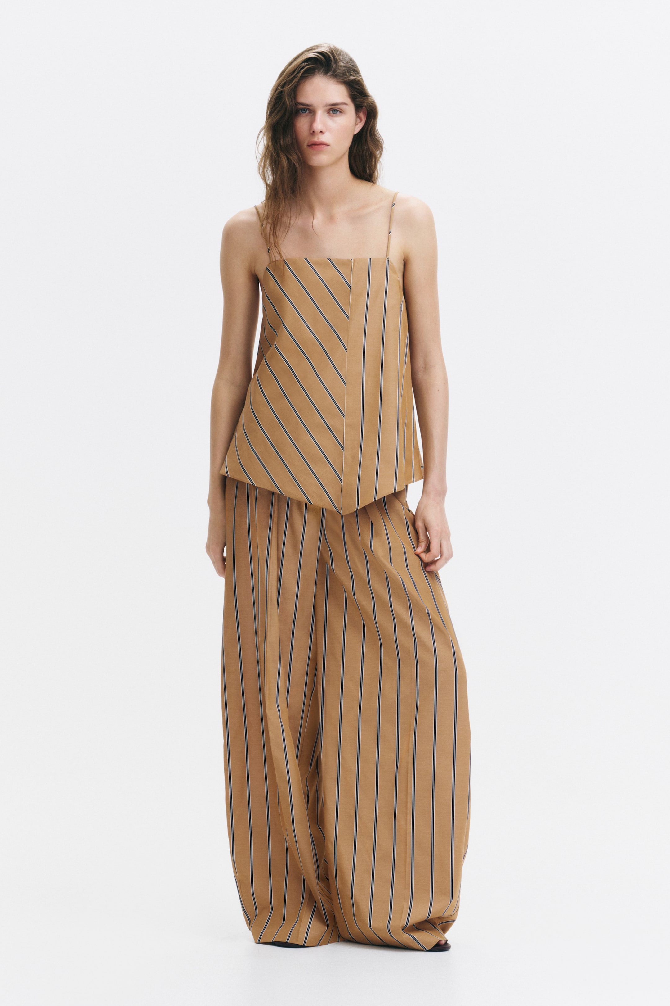 View larger image: The model is showcasing a relaxed-fit top crafted from tan woven fabric adorned with black stripes. It features a square neckline, delicate spaghetti shoulder straps, an A-line silhouette, and a subtly asymmetric pointed hem at the front. The top boasts a pattern of vertical stripes complemented by a diagonal striped panel, paired effortlessly with matching wide-leg pants.