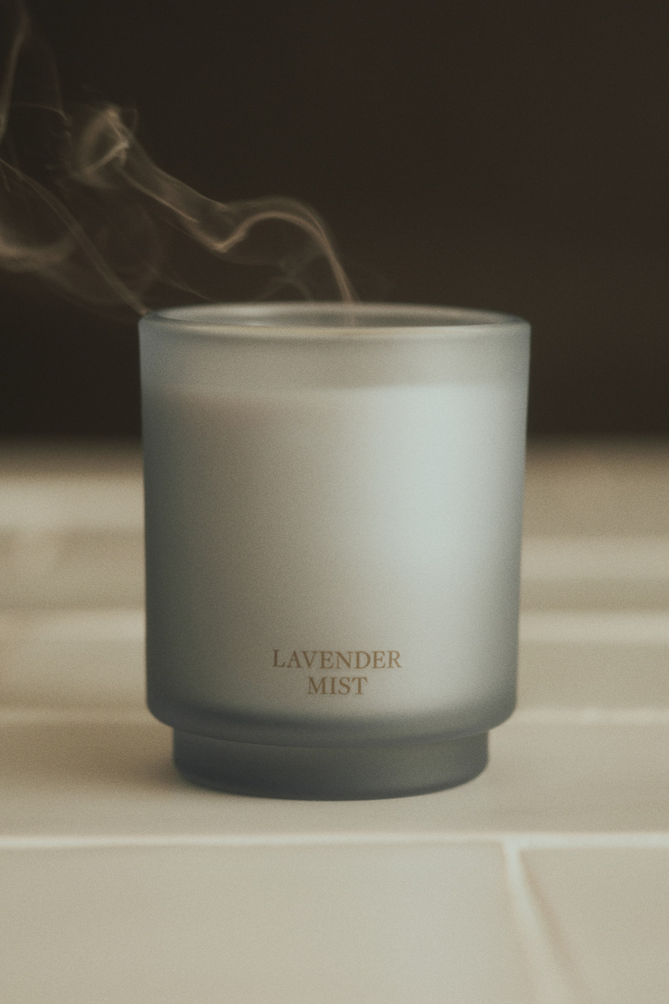 Grotere afbeelding bekijken: Een lavendelmistkaars in een mat, gedempt blauwgrijs glazen potje staat rechtop en toont gouden letters aan de voorkant met de tekst "LAVENDER MIST". Het potje heeft een cilindervormig lichaam met een iets smallere, getrapte voet, terwijl er een dun spoor rook opstijgt vanaf de bovenkant van de kaars.
