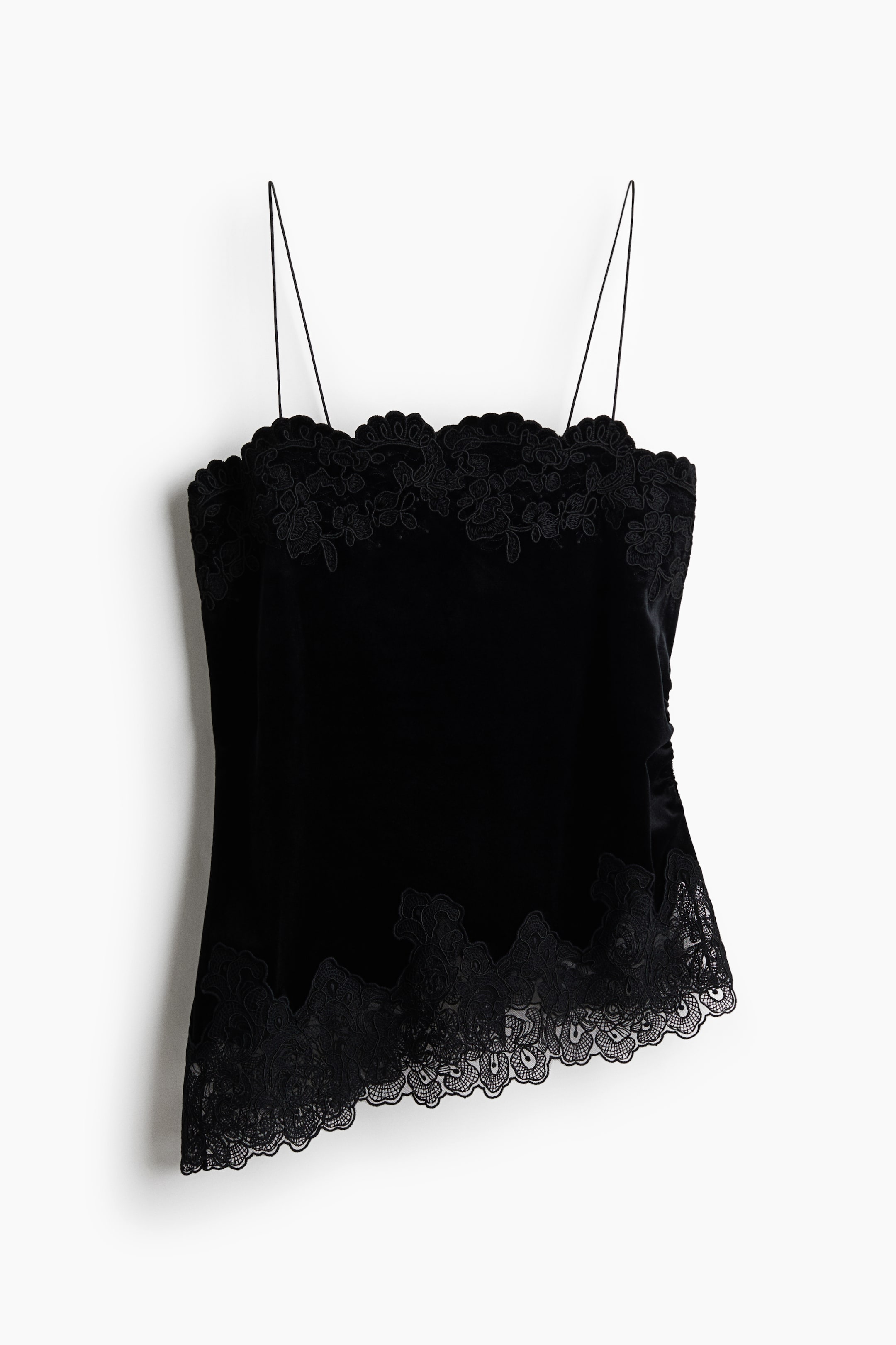 Visualizza immagine più grande: Un top camisole in velluto nero è appeso, mostrando il design frontale con spalline spaghetti ultra sottili e un raffinato bordo in pizzo smerlato lungo la scollatura dritta. Il top presenta un corpo in tessuto morbido e avvolgente che sfocia in un ampio bordo abbinato in pizzo smerlato lungo l’orlo asimmetrico.