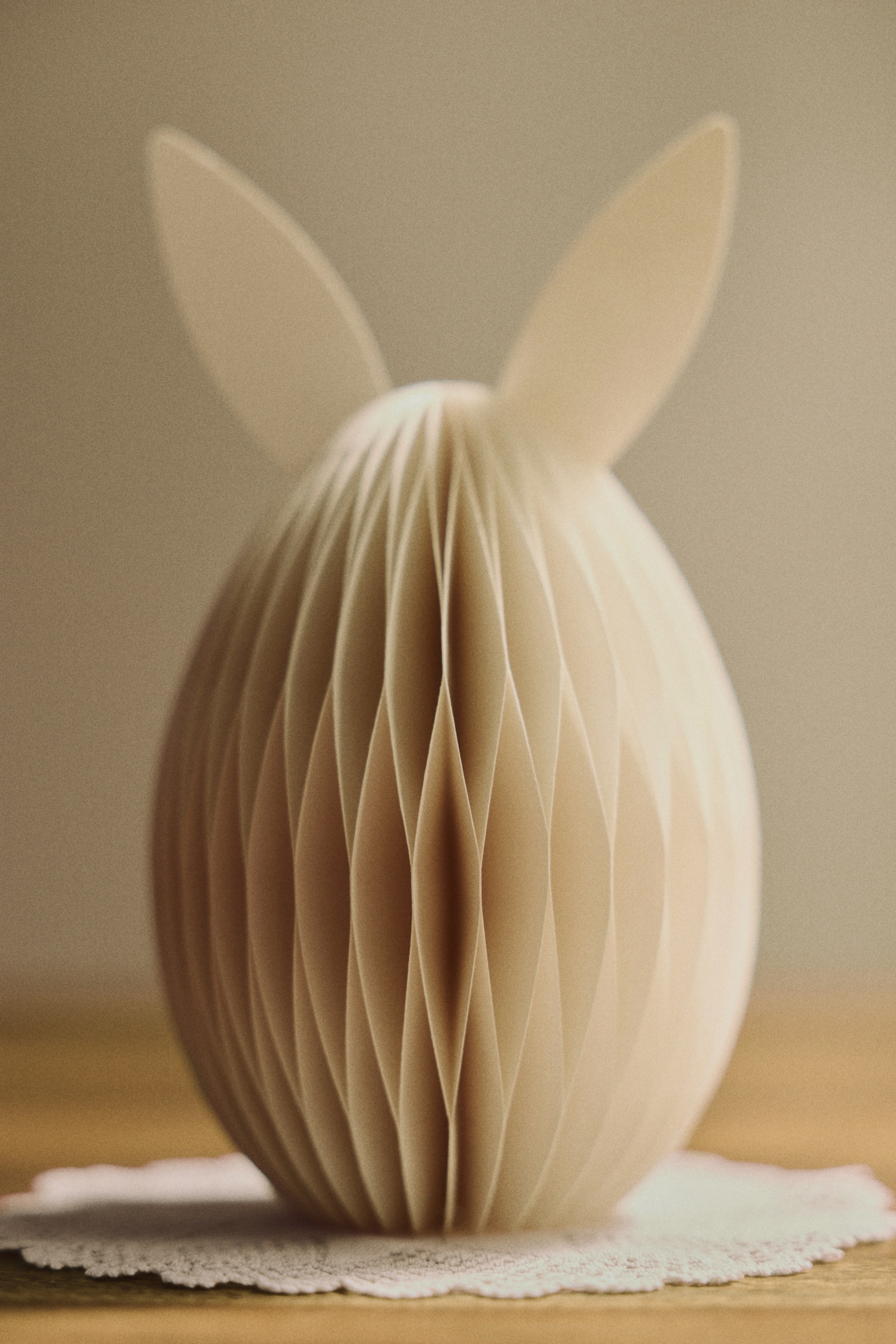 Grotere afbeelding bekijken: Een paasdecoratie toont een eivorm aan de voorkant, vervaardigd uit crèmekleurig honingraatpapier. Speelse konijnenoren steken uit de bovenkant.