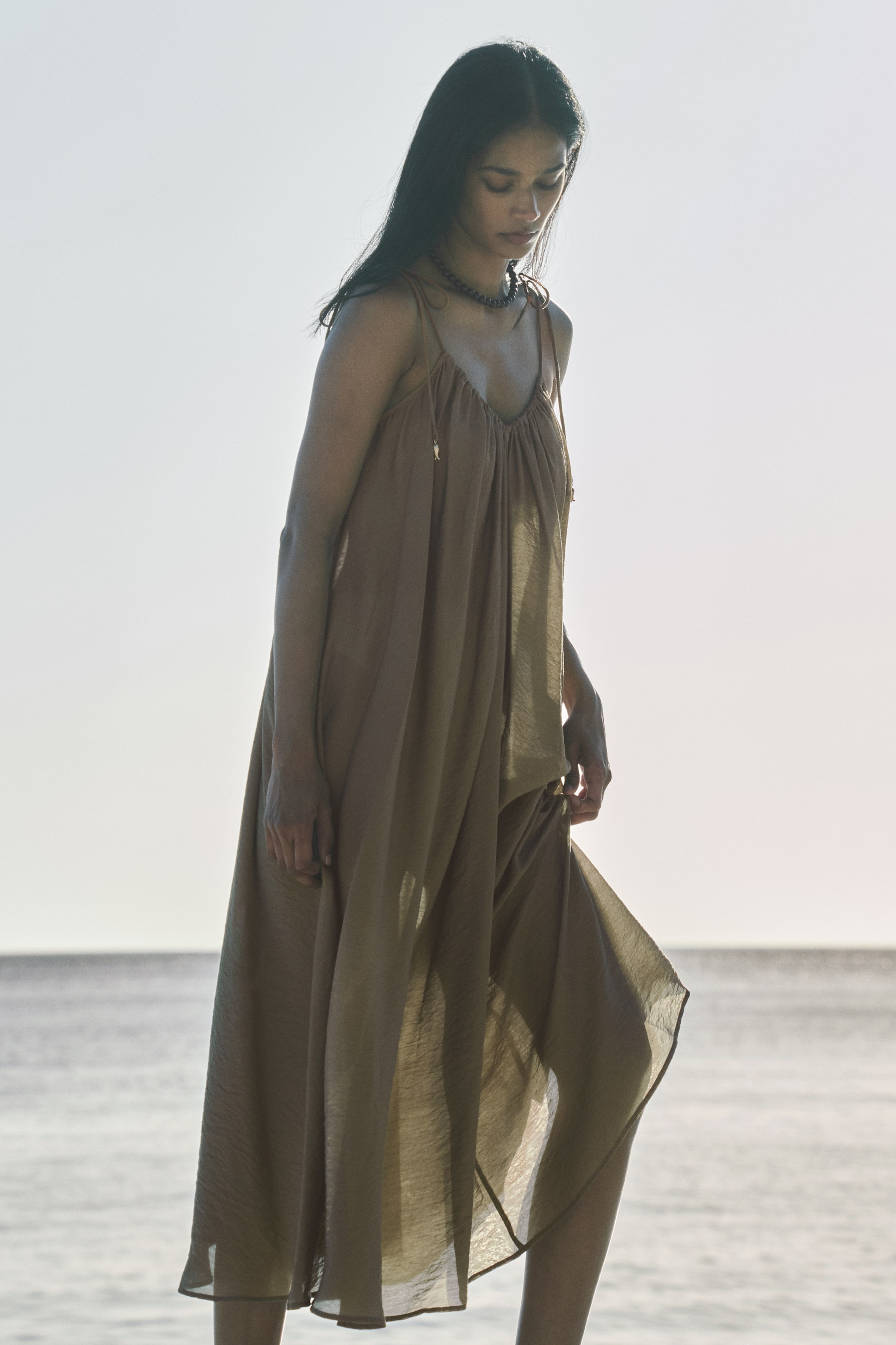 View larger image: The model is showcasing a long, loose-fit dress crafted from a gently crinkled weave. It features a V-neckline with tie-top spaghetti shoulder straps that elegantly tunnel through the front, creating gathers that give rise to a voluminous A-line silhouette. The model is softly holding the fabric of the dress, highlighting its flow and texture.