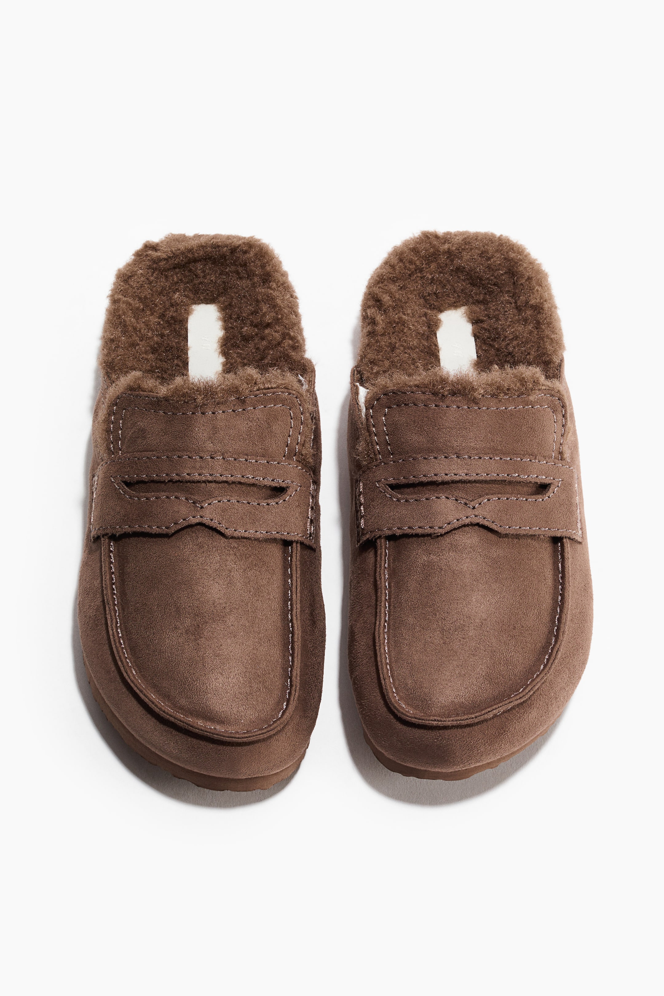 View larger image: A pair of brown loafer-style slippers is facing the back, revealing a fluffy brown interior lining. They feature a suede-like exterior with a strap across the vamp and visible stitching.