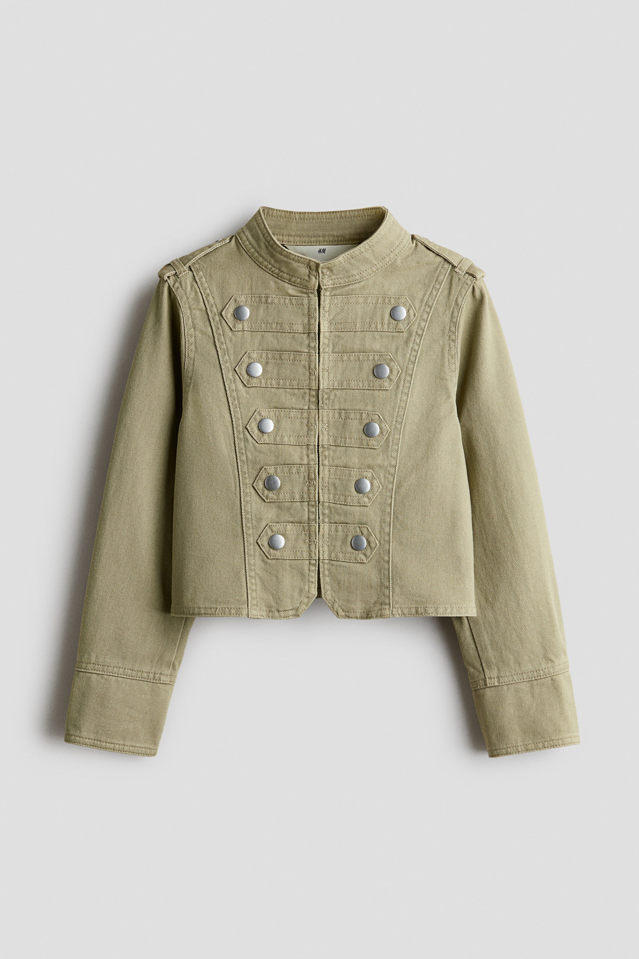 Agrandir l'image: Cette veste courte en sergé de coton vert olive présente, de face, un design d’inspiration militaire, avec un petit col montant et une fermeture frontale dissimulée. Elle affiche cinq pattes cousues à clous avec des clous finition argent de chaque côté de la poitrine, ainsi que des pattes d’épaule et des manches longues à larges poignets.