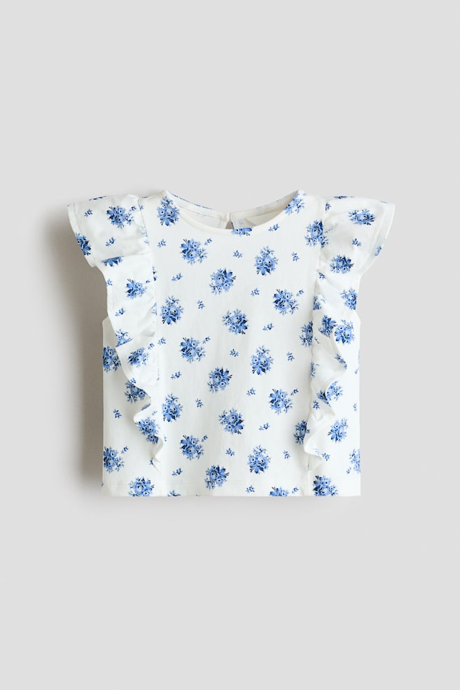 Ein weißes Oberteil präsentiert einen blauen Blumenprint mit verstreuten kleinen Blumenclustern und einzelnen Blüten, ausgestattet mit gerüschten Kappärmeln und einem runden Halsausschnitt. Das Oberteil wird von vorne gezeigt, mit einer kleinen Schlüssellochöffnung und einem Knopfverschluss, der am hinteren Halsausschnitt sichtbar ist. - 1