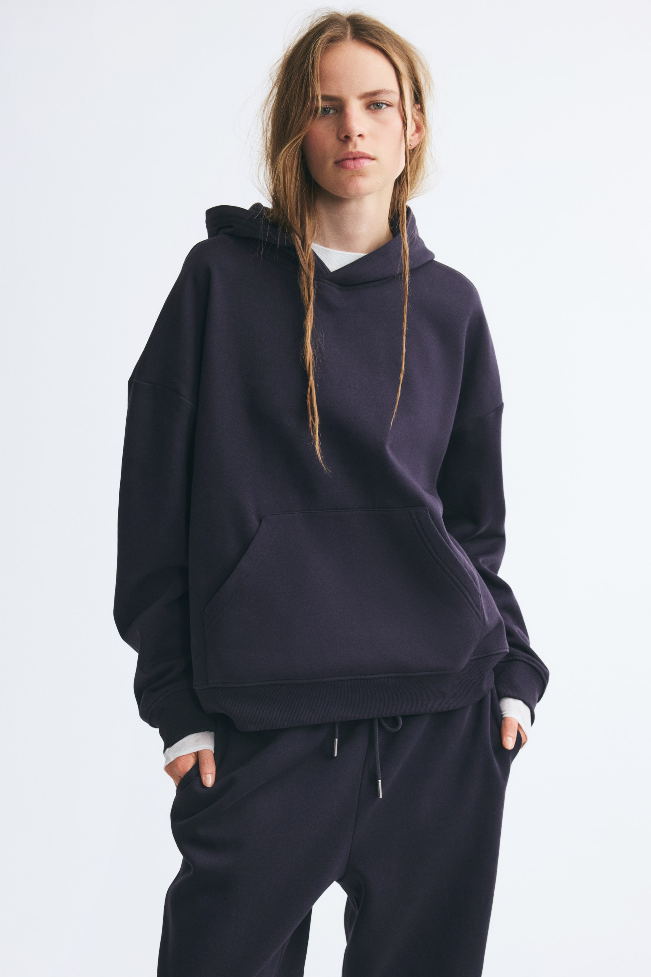 Se større billede: En mørkelilla hoodie i sweatstof med sænkede skuldre, kængurulomme og ribkanter ved ærmerne bæres af modellen. Hoodien er stylet over en hvid top og kombineret med matchende mørkelilla sweatpants.