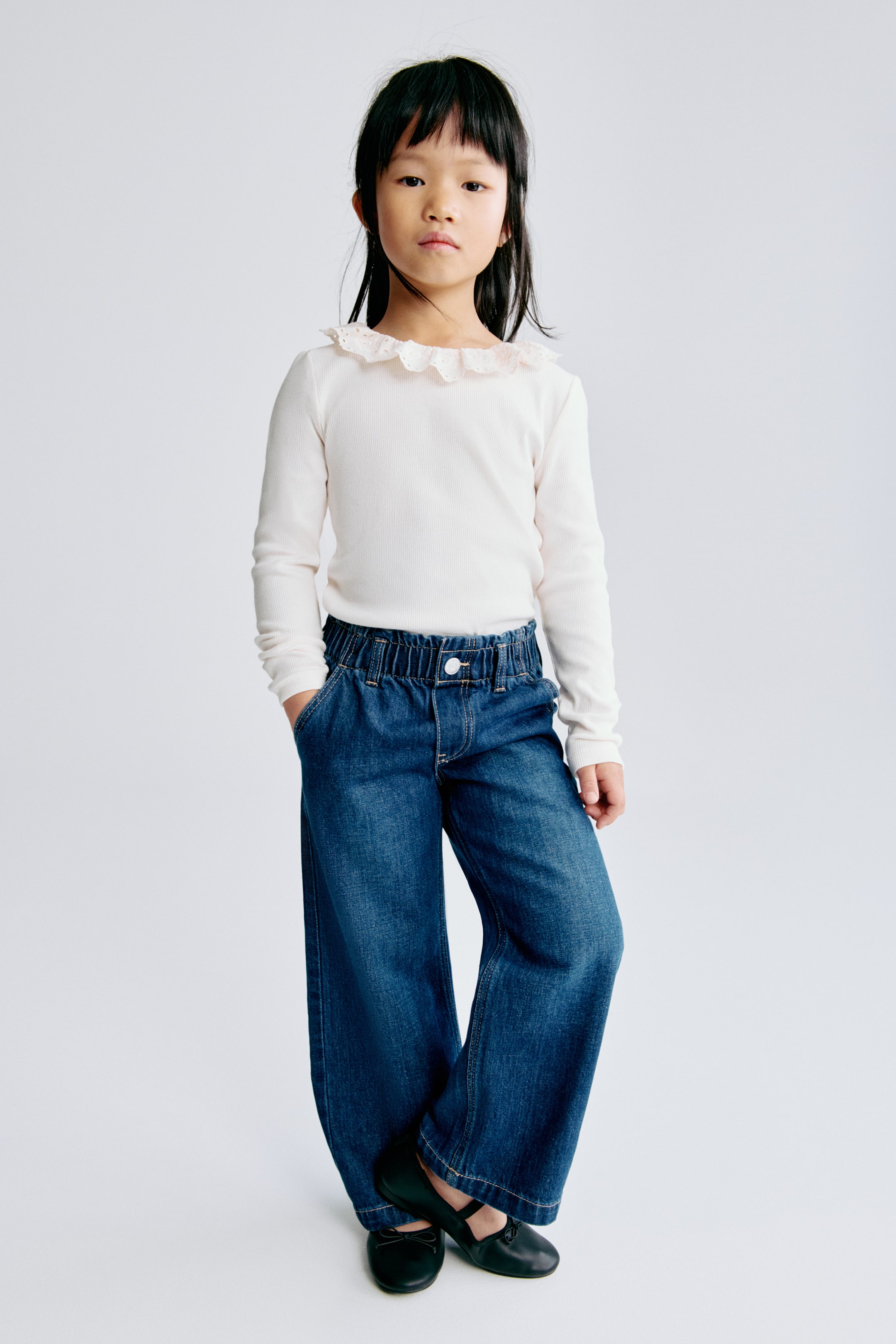 View larger image: An off-white ribbed long-sleeved top with a ruffled lace collar is being worn, paired with dark blue wide-leg jeans featuring an elasticized paperbag waist and visible side pockets.