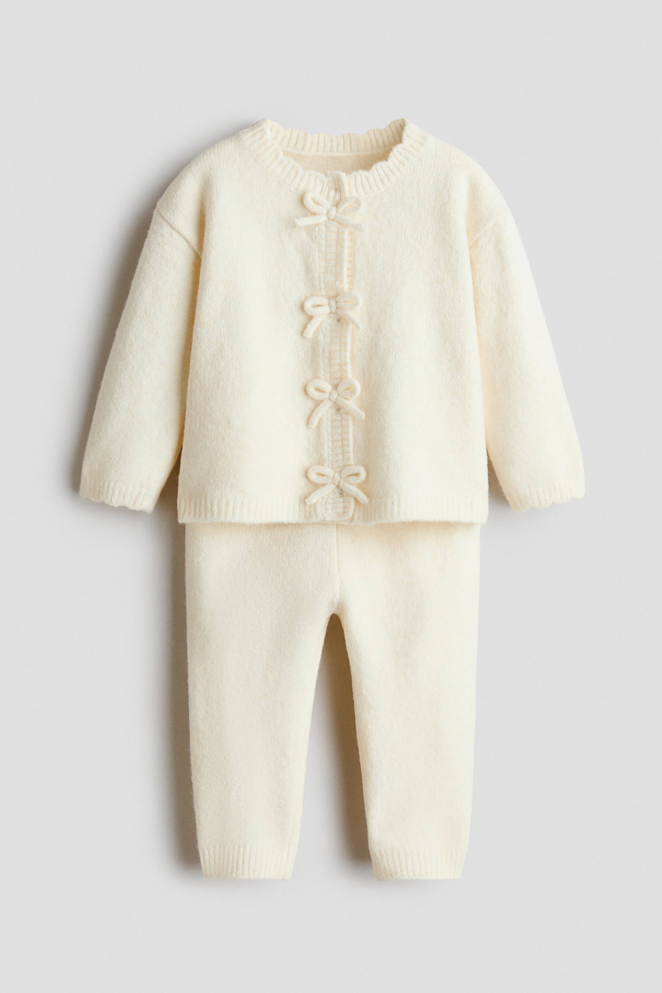 View larger image: A cream fuzzy knitted two-piece baby outfit is on display. The cardigan features a scalloped neckline and three decorative bows down the front placket, with ribbed cuffs and hem. Matching knitted pants feature ribbed ankle cuffs.