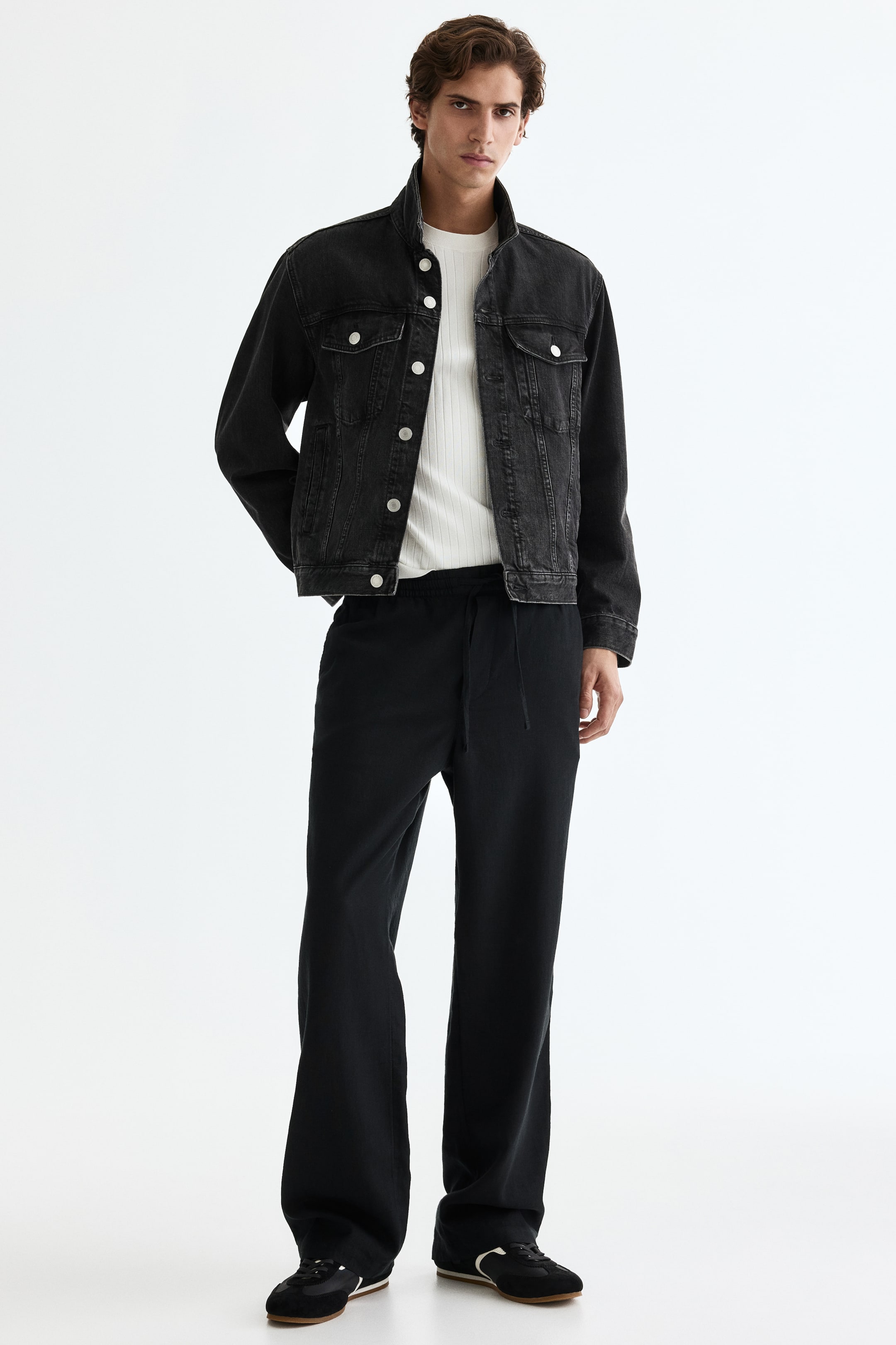 Größeres Bild anzeigen: Eine schwarze Jeansjacke wird von vorne präsentiert, mit einer entspannten Passform, einem Stehkragen und einem Knopfverschluss mit silberfarbenen Knöpfen. Sie zeigt zwei Brusttaschen mit Klappen und Knöpfen sowie zwei seitliche Leistentaschen und wird über einem weißen gerippten Oberteil getragen.