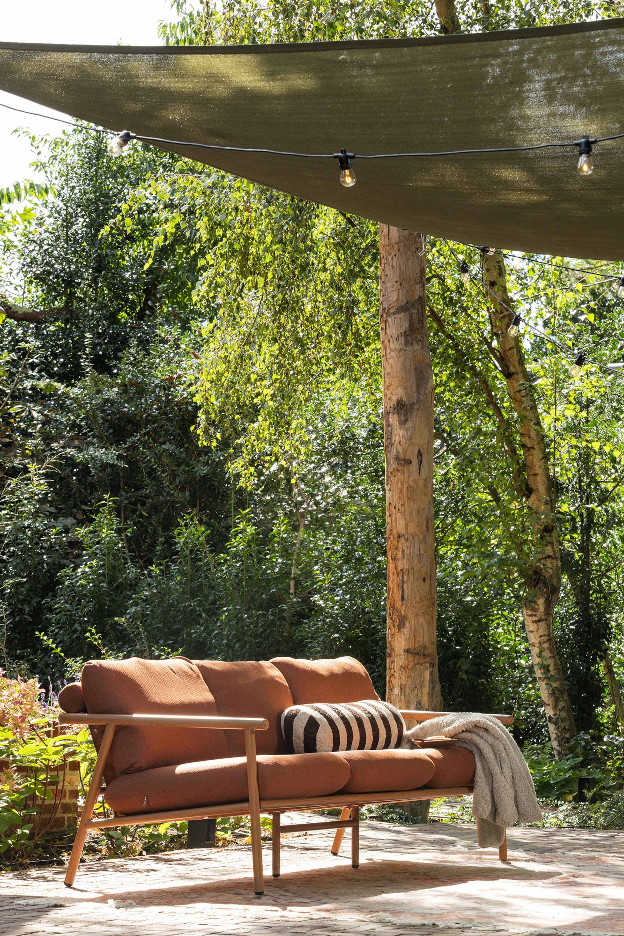 Größeres Bild anzeigen: Das Zweisitzer-Sofa präsentiert sich mit einem hell getönten Holzrahmen und üppigen, terrakottafarbenen Kissen. Es zeigt seine Vorder- und rechte Seite, mit einem schwarz-weiß gestreiften Nackenpolsterkissen, das an einem Ende liegt, und einer hellgrauen Überwurfdecke, die über die andere Armlehne und den Sitz drapiert ist.