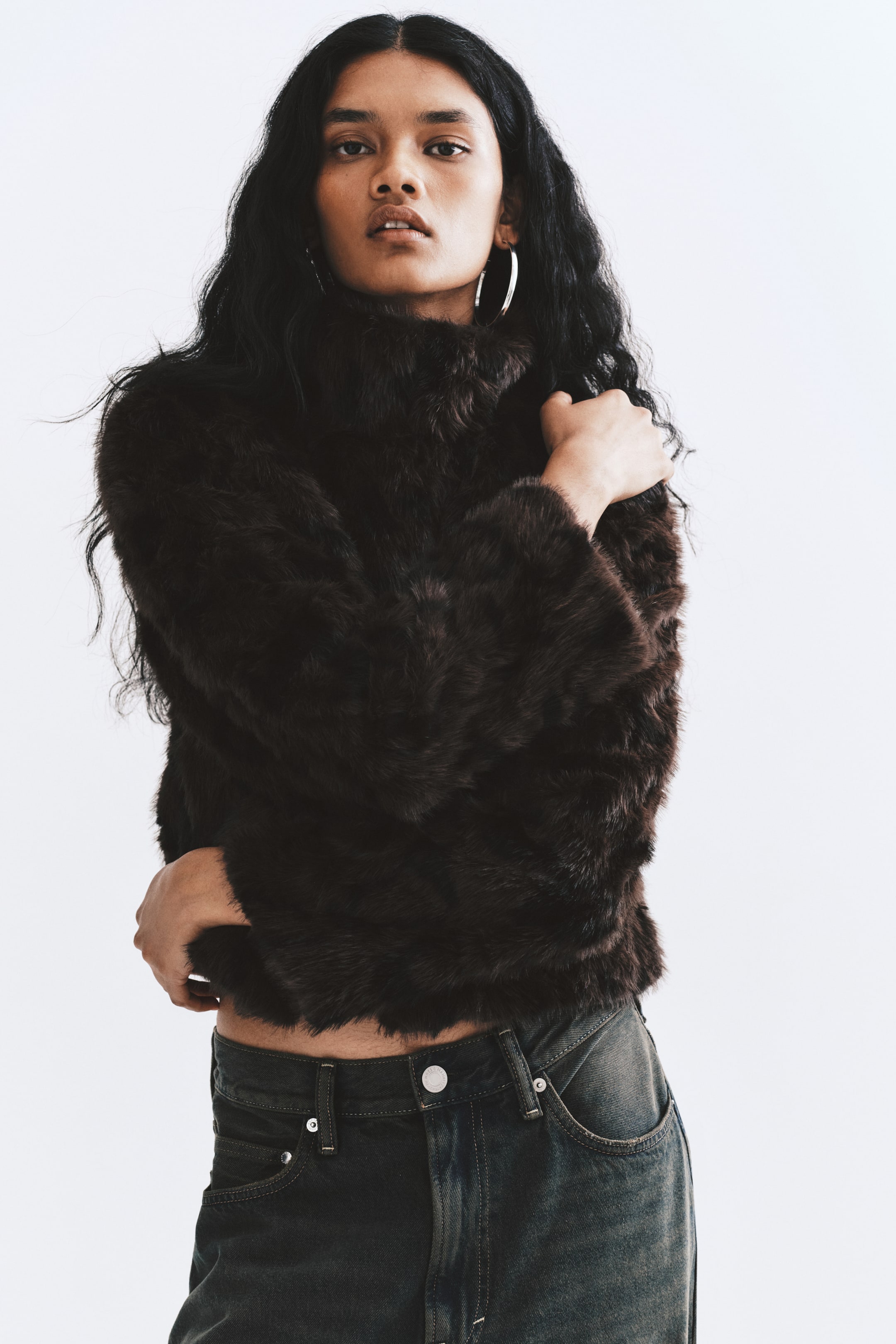 View larger image: A short jacket in dark brown fluffy fabric is being worn by the model, who is posing with their arms crossed over their chest. The jacket features a high, double-layered funnel collar and is paired with dark grey jeans.