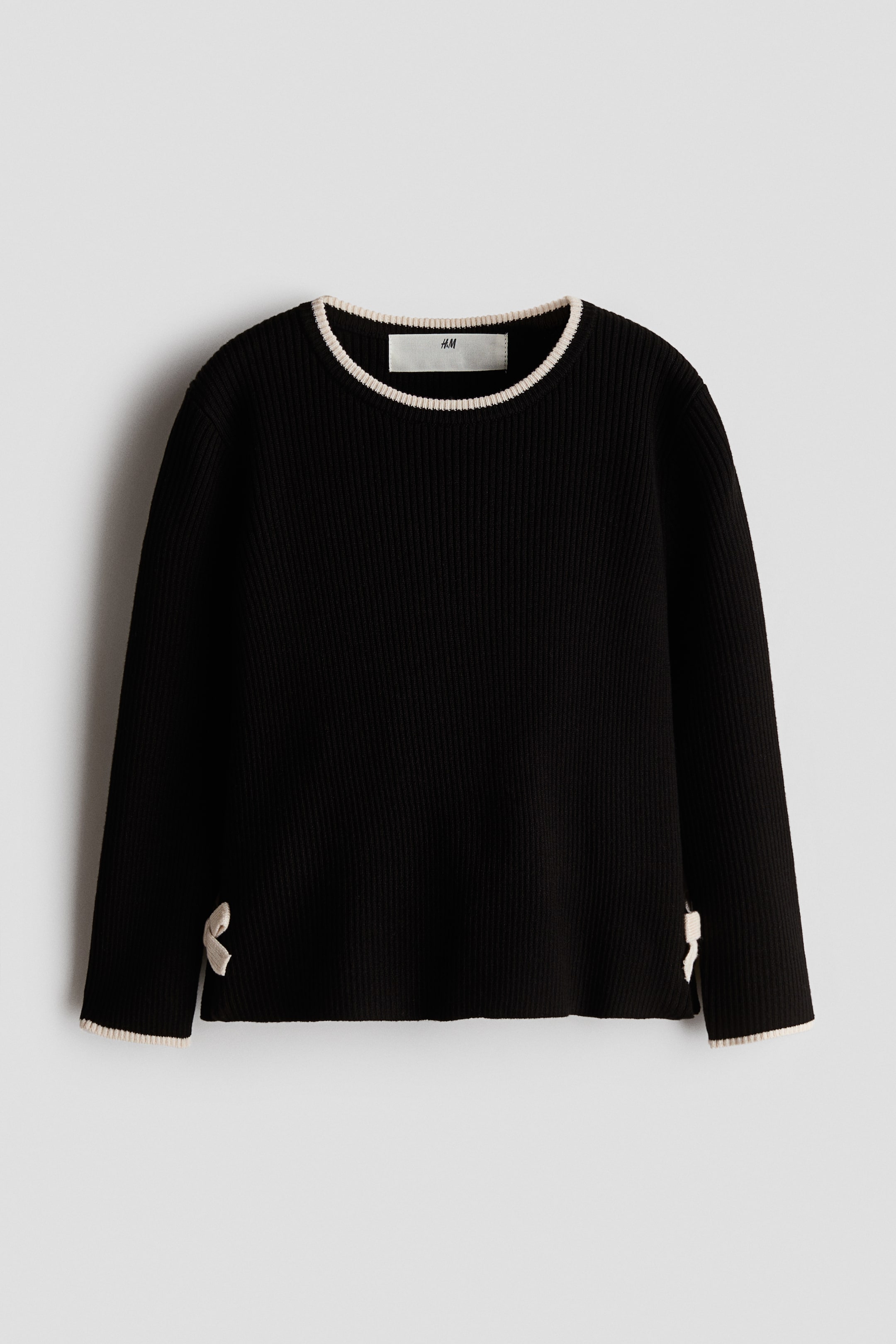 View larger image: A black ribbed knit sweater is shown with a round neck and cream-coloured trim, with a white H&M tag visible at the back of the neck. Long sleeves finish with cream-coloured trimmed cuffs, and the straight hem also features a matching trim, along with side slits tied with small cream-coloured bows.