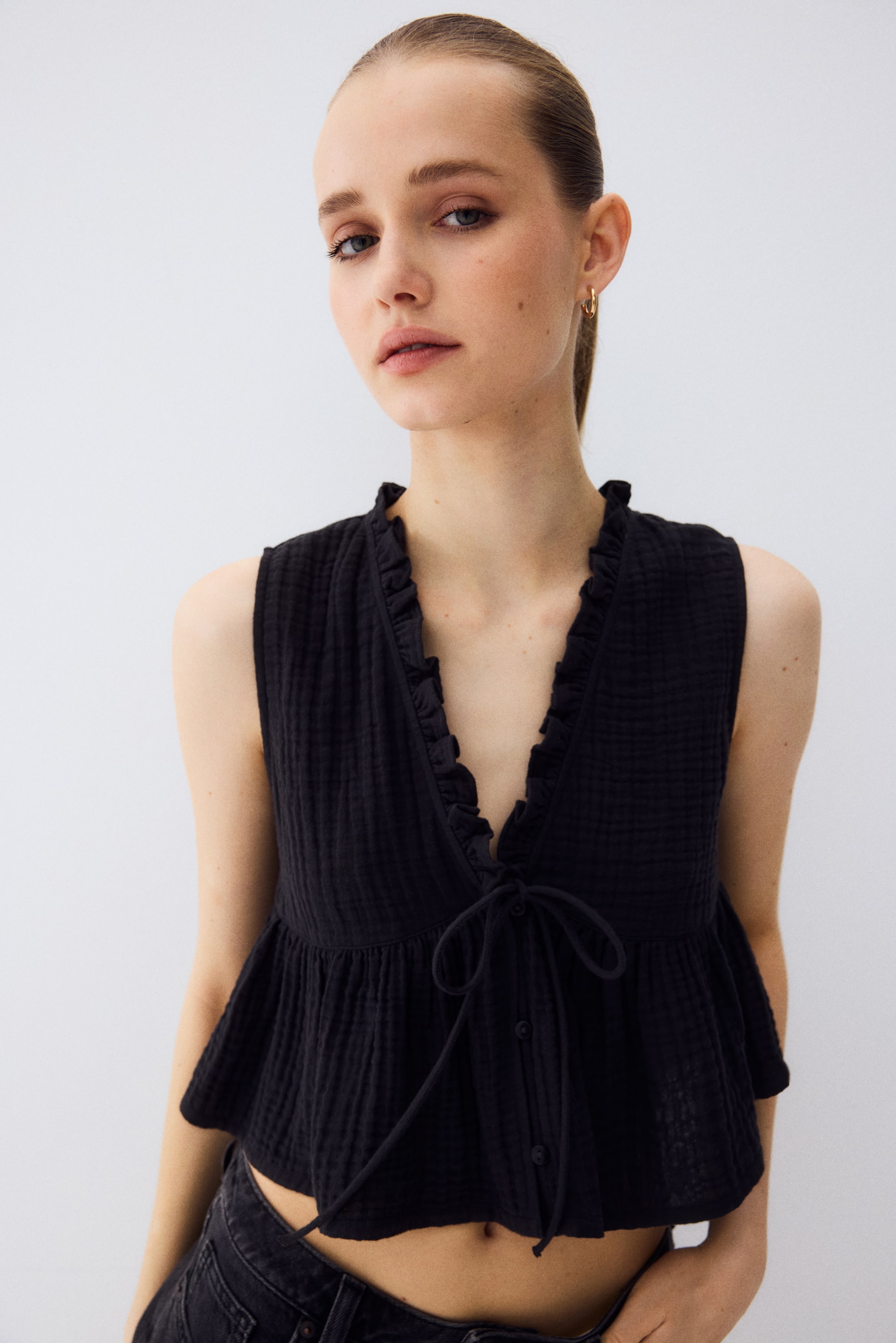 View larger image: A black sleeveless top showcases a crinkled texture, adorned with a ruffled V-neckline and a ruffled neck edge. It features a tie-front closure over buttons, crafted in a peplum crop style, slightly angled towards the viewer's right.