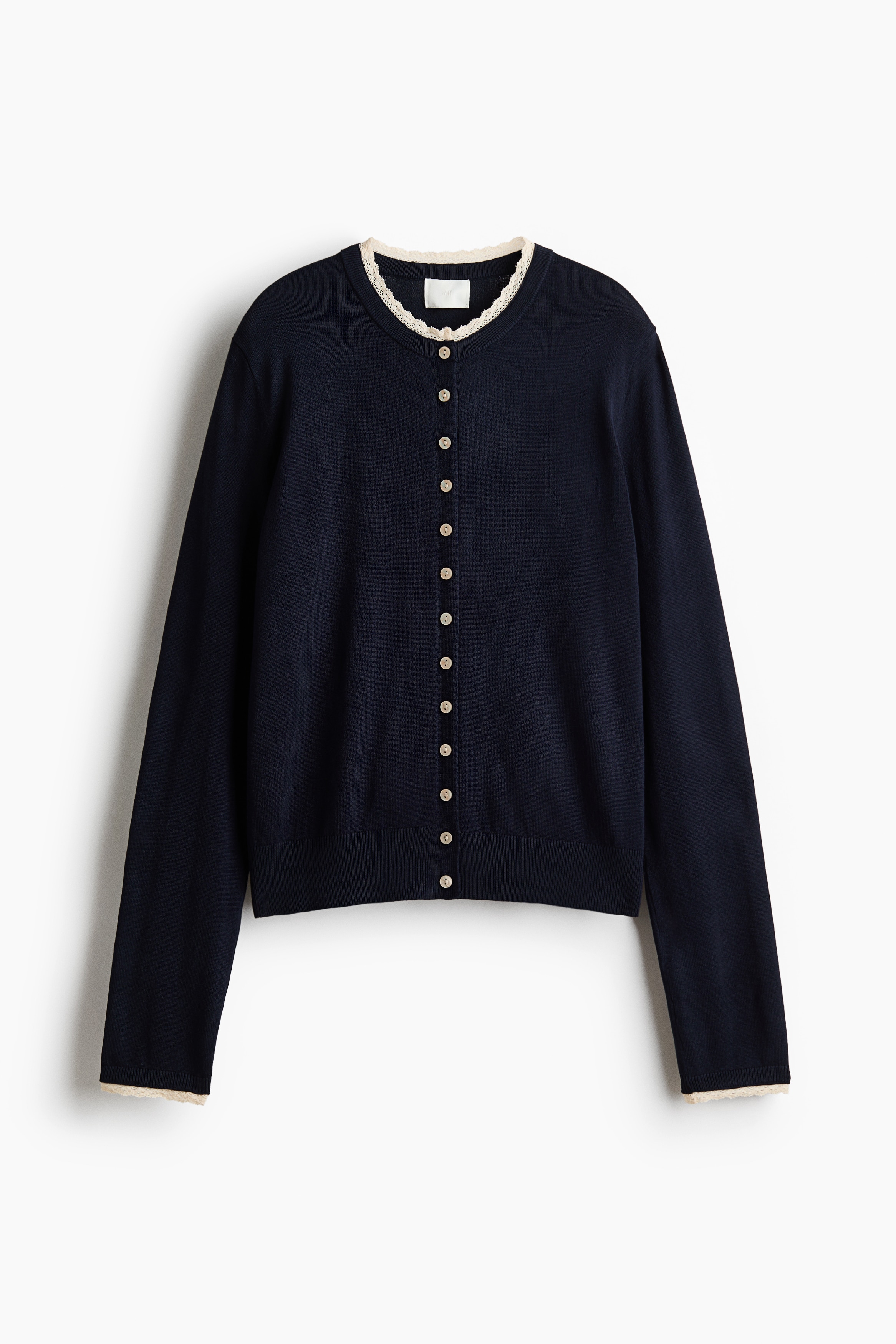 A dark navy blue, long-sleeve knit cardigan is shown from the front, layered over a thin white long-sleeve top. The cardigan features a round neckline with cream-coloured lace trim, small light buttons down the front, and ribbed detailing on the hem and cuffs, which are also trimmed with cream lace.