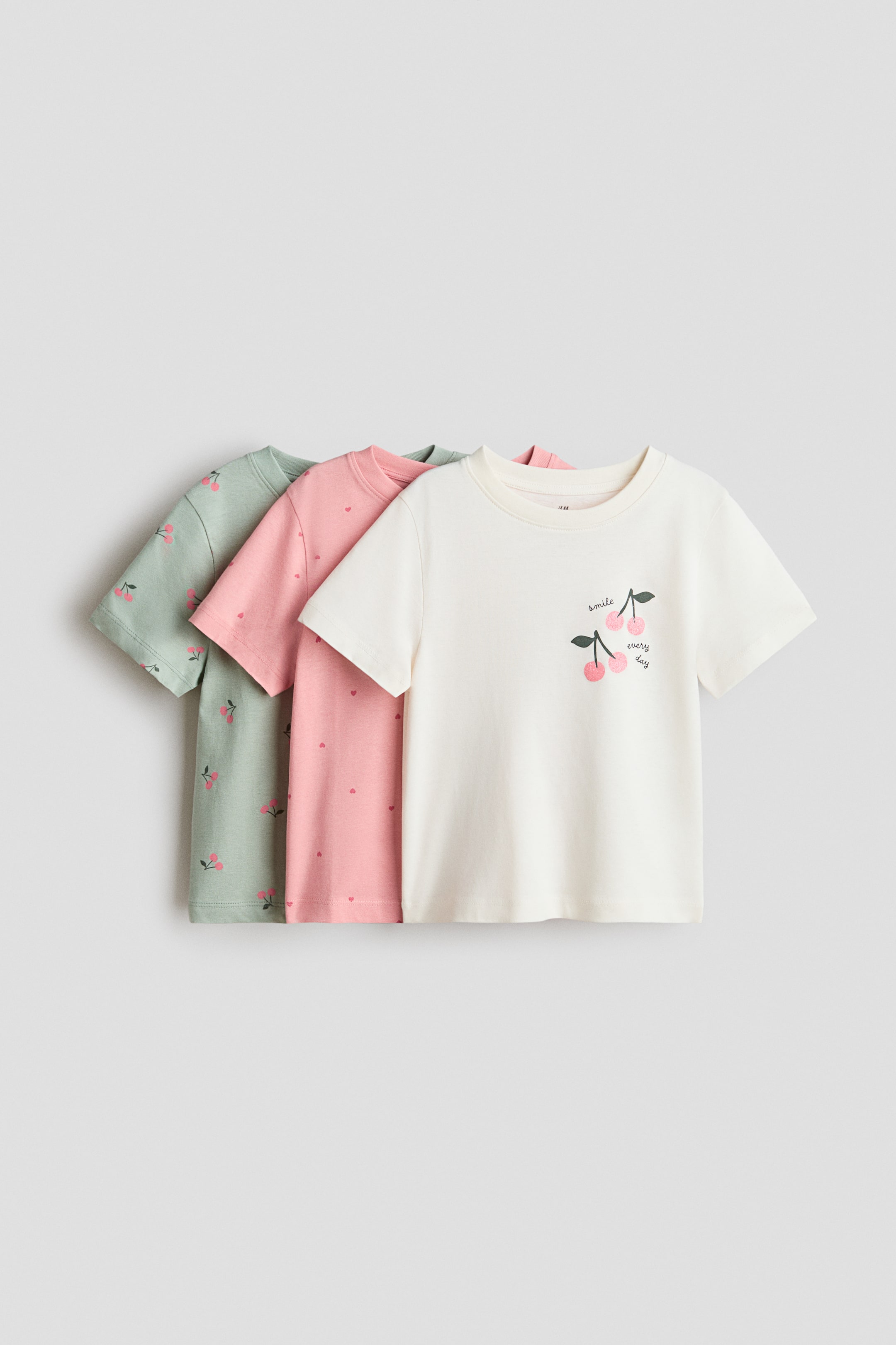 View larger image: A set of three short-sleeve crewneck T-shirts is shown from the FRONT, layered from front to back. The front off-white T-shirt features a cherry graphic with the text “smile every day” on the left chest. The middle pink T-shirt displays an allover heart pattern, and the back light green T-shirt shows an allover cherry pattern.