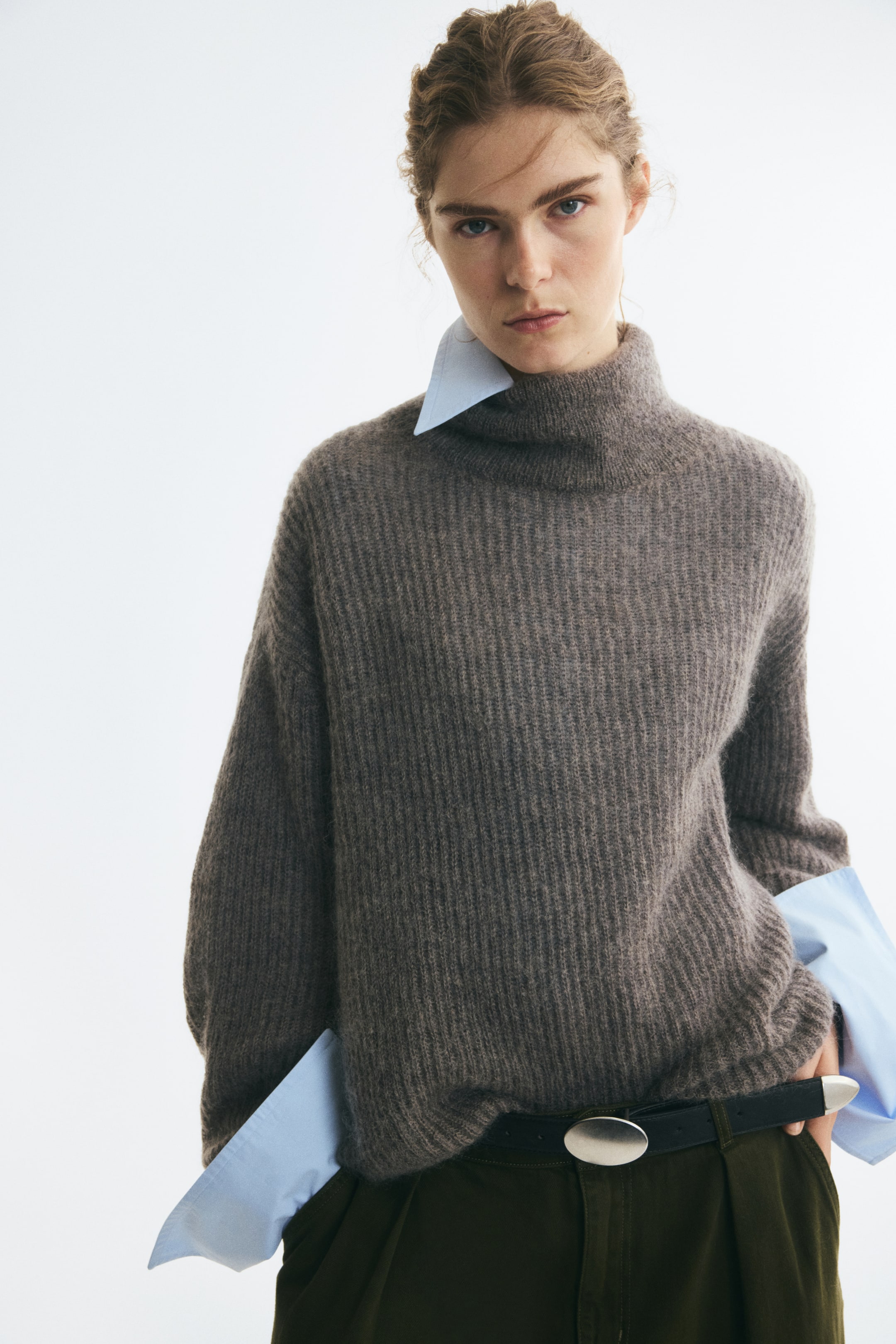 Ver imagen más grande: Una modelo lleva un jersey oversize de mezcla de mohair con textura canalé en tono topo-marrón, con cuello alto, hombros caídos y mangas largas. El jersey se combina sobre una camisa de cuello azul claro, con los puños asomando por debajo de las mangas, y se complementa con un pantalón verde oscuro.