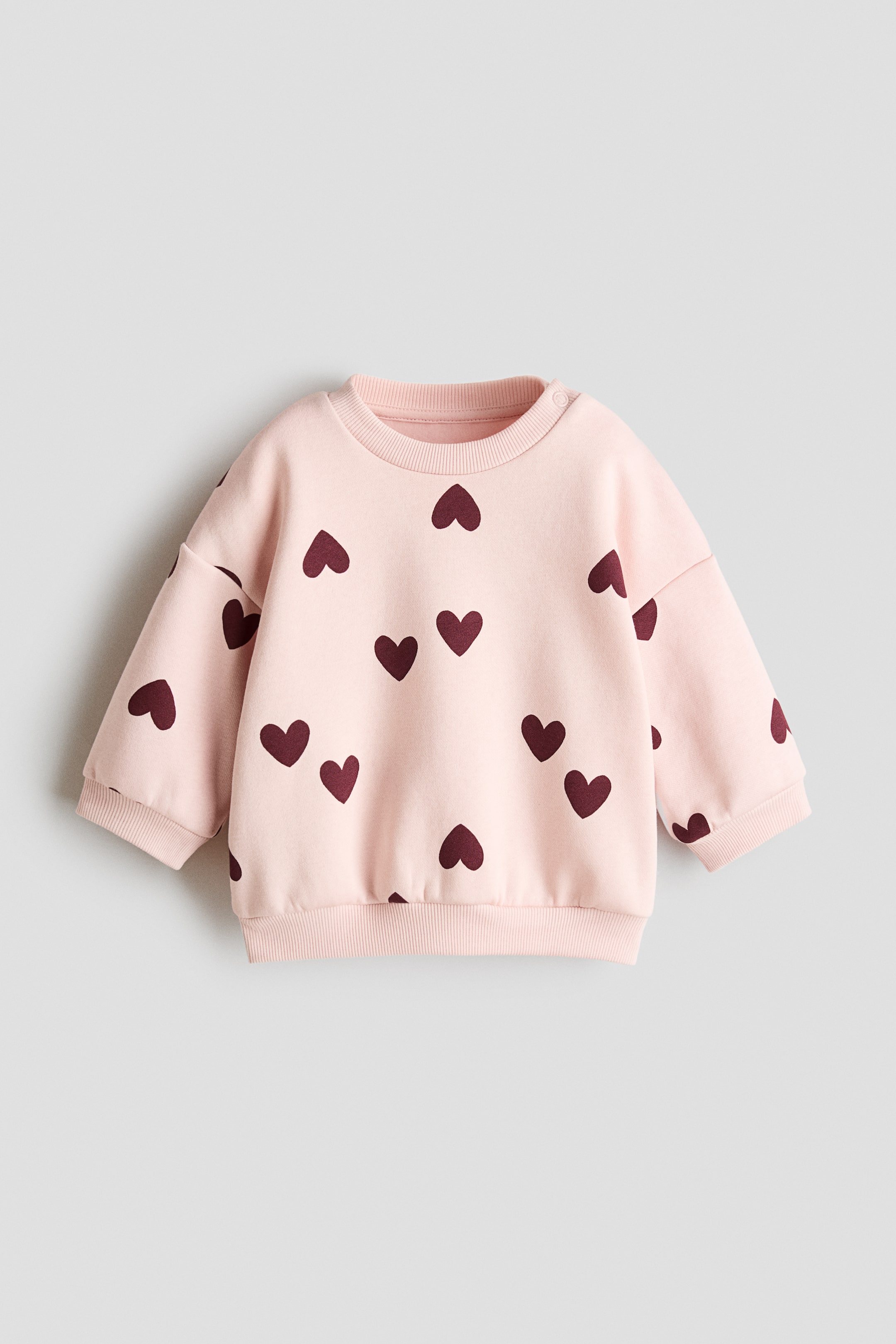 View larger image: A light pink baby sweatshirt featuring a pattern of dark red hearts across the front. It is designed with a ribbed crew neck, ribbed cuffs, a ribbed hem, and two snap buttons on the right shoulder.