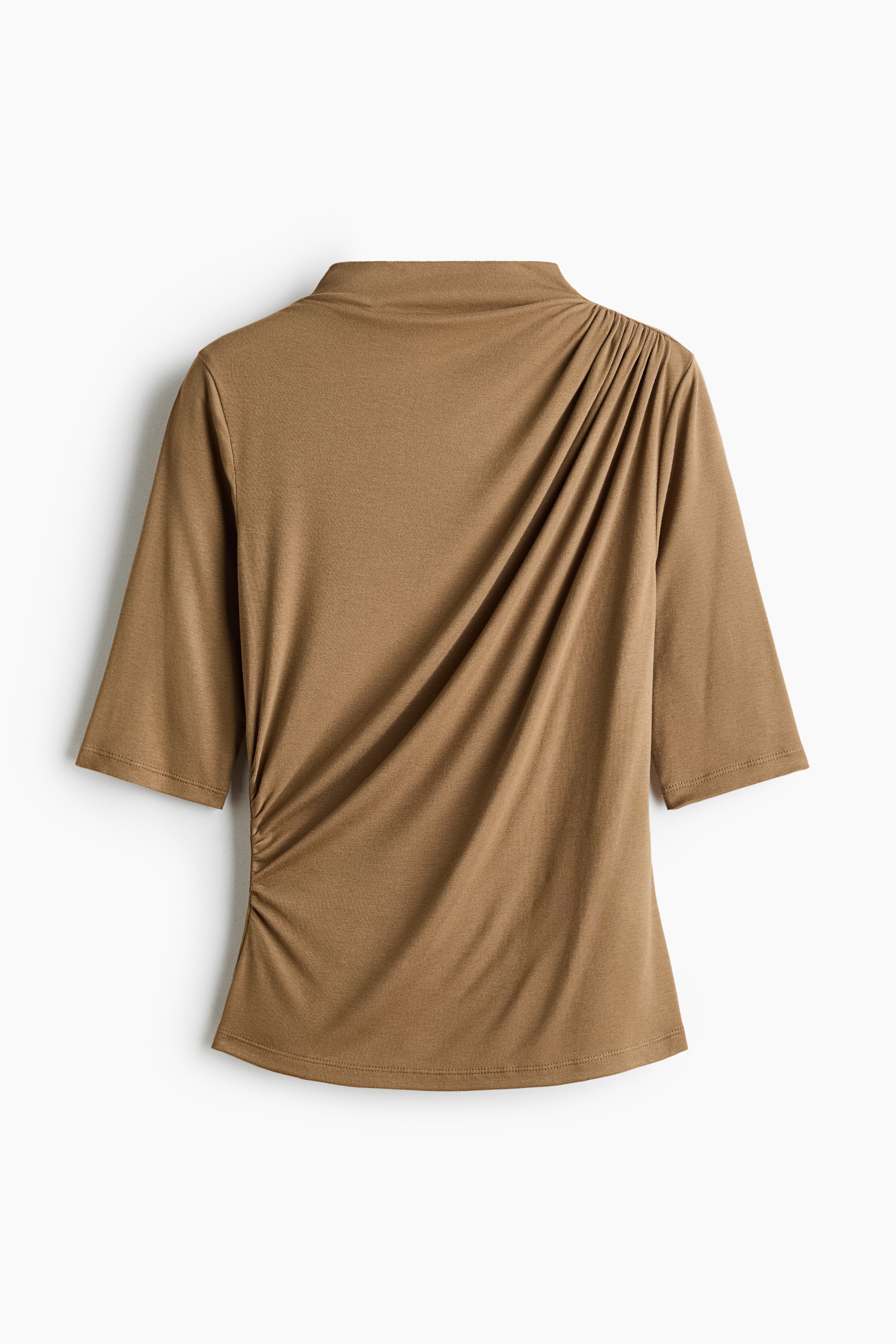 La modelo lleva una blusa ajustada de jersey color café claro, con cuello alto y mangas a la altura del codo. La prenda presenta un detalle fruncido asimétrico que cae sobre el pecho y el torso, combinada con jeans de mezclilla de lavado claro.