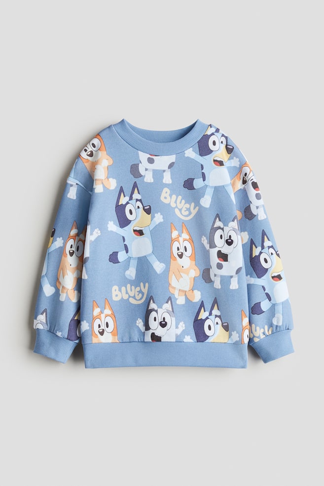 Ein hellblaues Oberteil aus bedrucktem Sweatshirtstoff wird in einer FRONT-Ansicht gezeigt, mit einem sich wiederholenden Muster verschiedener Cartoon-Hundefiguren und dem Wort "Bluey." Es verfügt über einen runden, gerippten Ausschnitt, überschnittene Schultern, lange Ärmel sowie gerippte Bündchen und einen gerippten Saum. - 1
