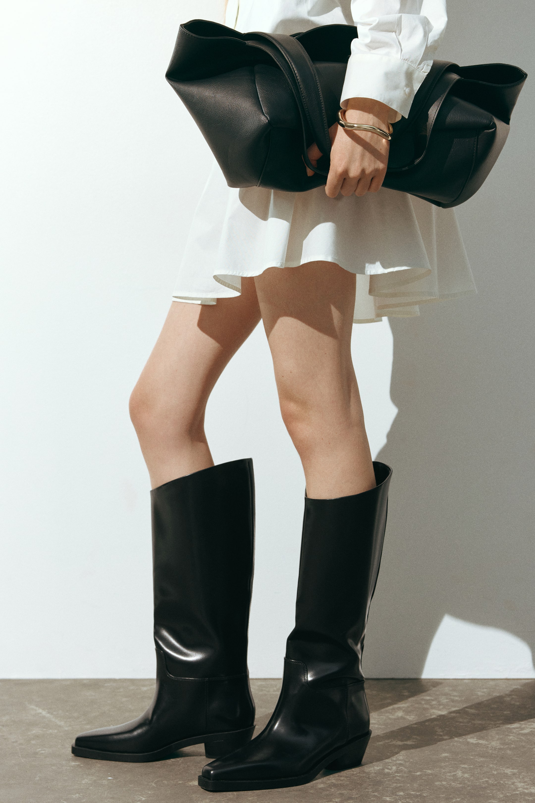 View larger image: The model is wearing knee-high boots in black coated fabric with chisel toes and low block heels. The boots are styled with a short white dress and a large black handbag.