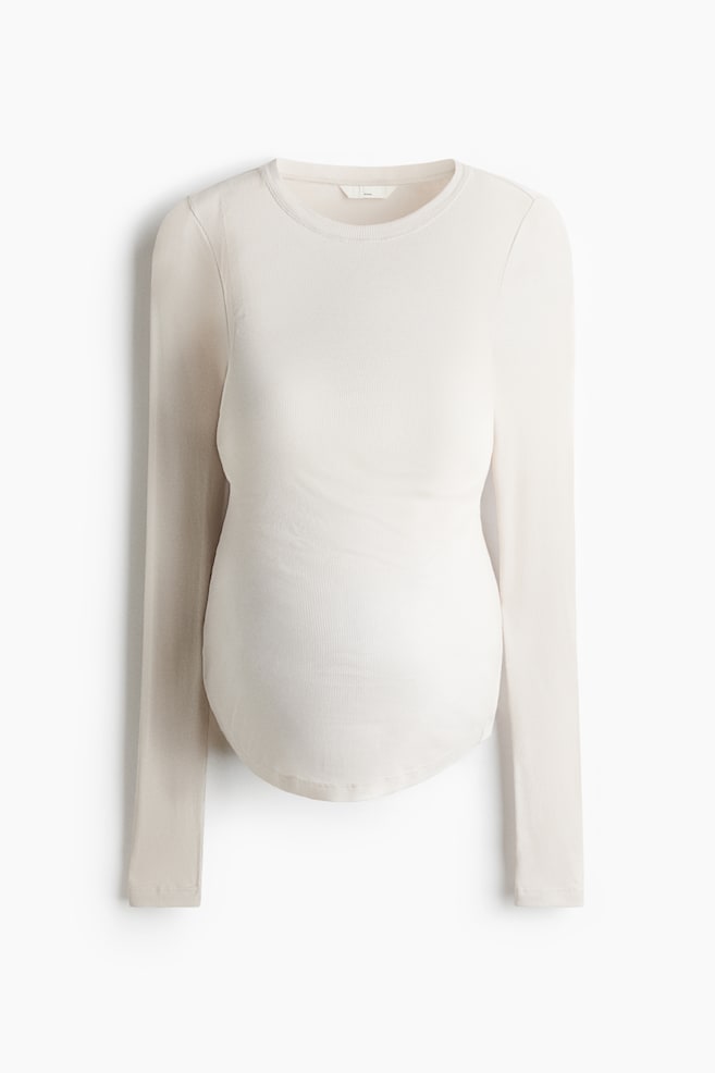 A light beige maternity top is shown from a front-facing angle, featuring a fine ribbed-knit texture, a crew neck, and long sleeves. The top has a fitted silhouette with a curved hemline that contours over a rounded pregnancy bump. - 1