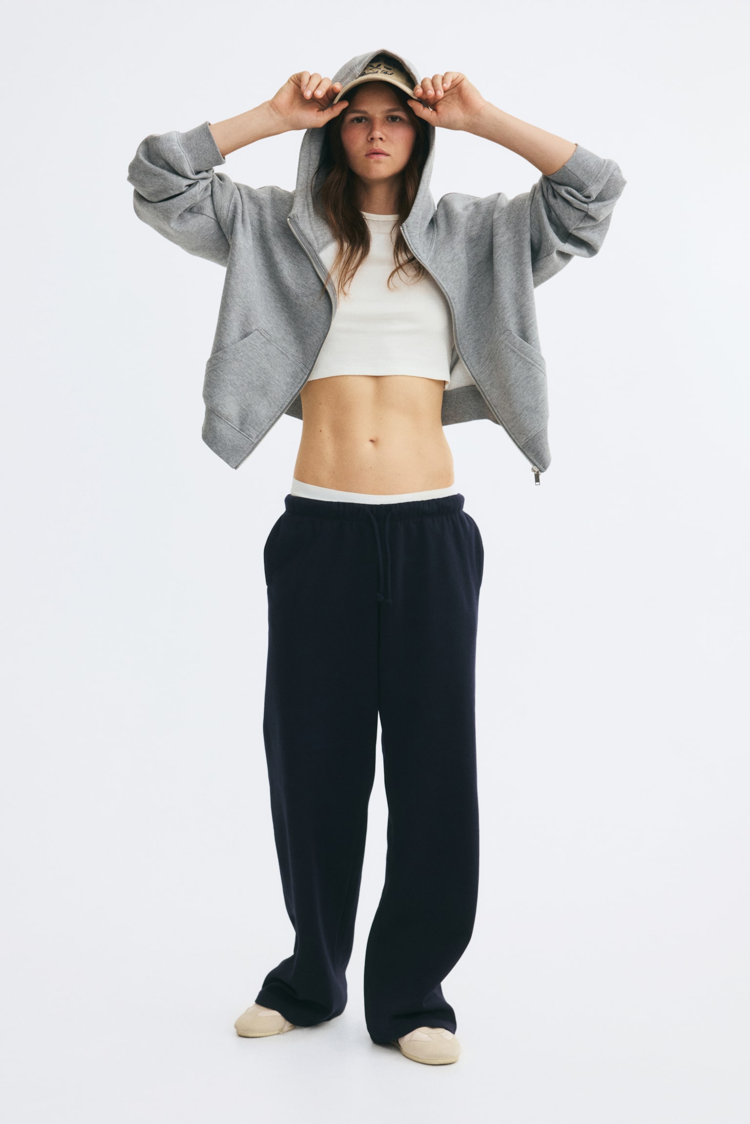 Wide-leg, loose-fit track pants in dark navy are being worn by the model, featuring a visible elasticated waistband with a drawstring. The pants are styled with a white cropped top and an open grey zip-up hoodie. - 1