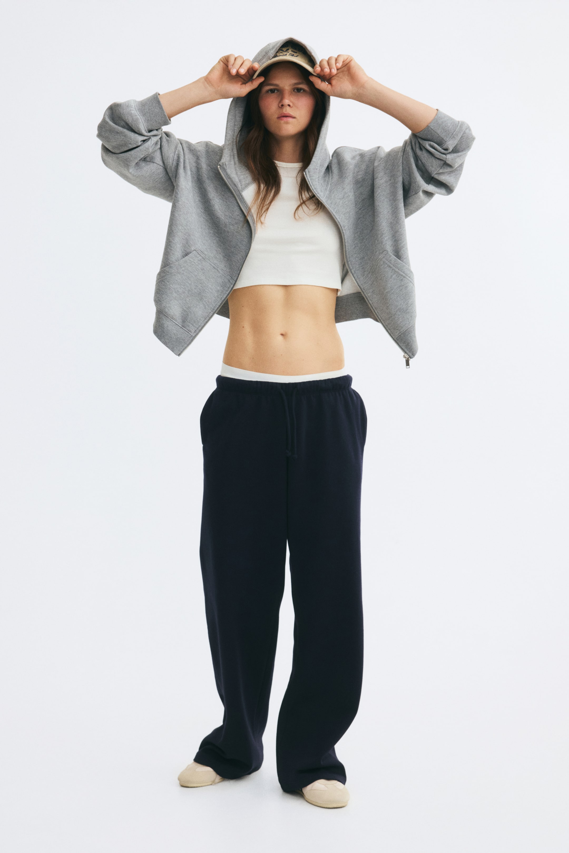 Größeres Bild anzeigen: Weit geschnittene, locker sitzende Jogginghose in Dunkelmarine, getragen vom Model, mit sichtbarem elastischem Bund und Kordelzug. Die Hose ist mit einem weißen Cropped-Top und einem offen getragenen grauen Zip-Hoodie kombiniert.