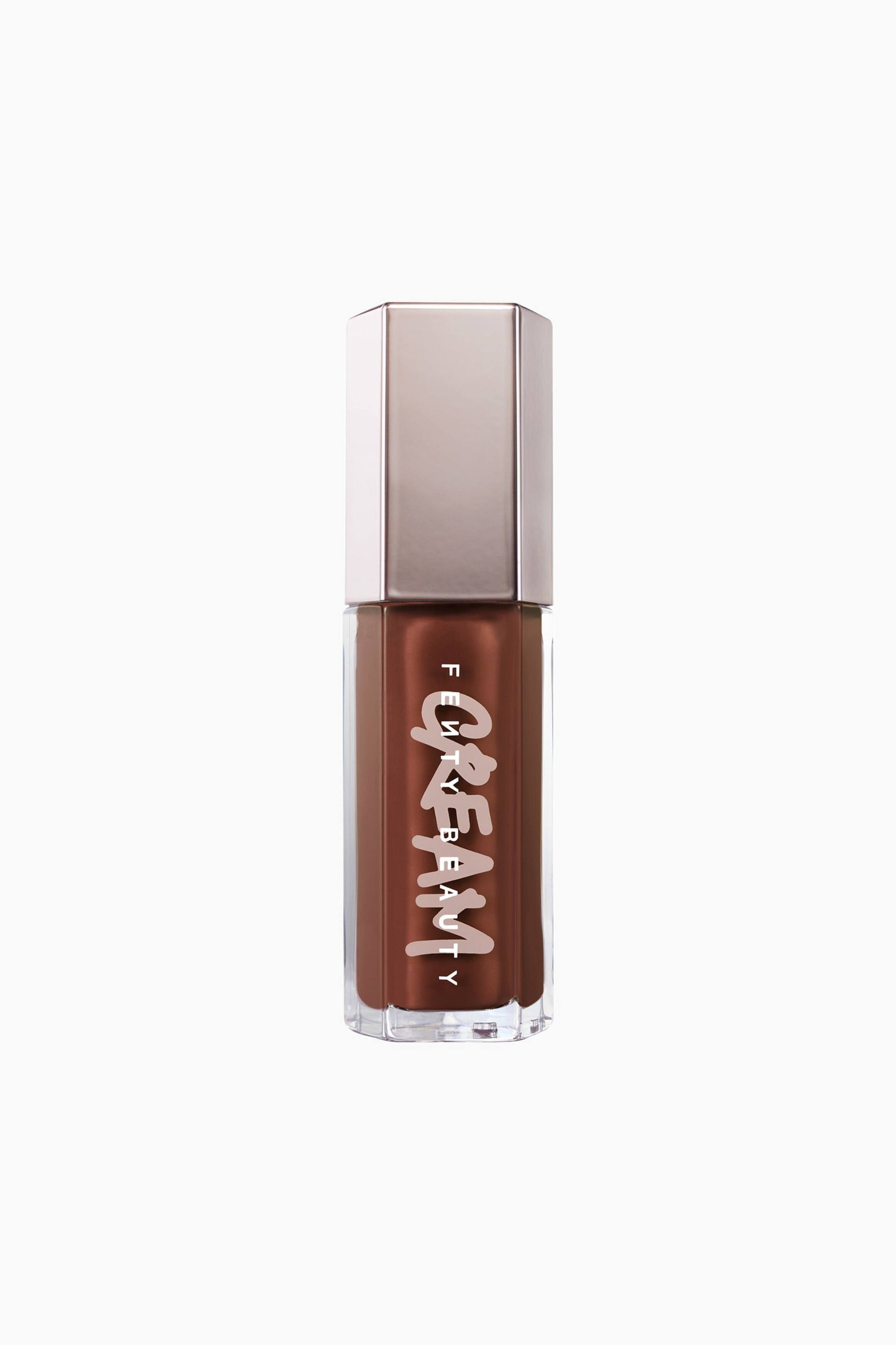Näytä suurempi kuva: Fenty Beauty -huulikiilto pullo on pystyasennossa, ja siinä näkyy kirkas kuusikulmainen tuubi, joka on täytetty kermaisella suklaanruskealla nesteellä. Tuubissa on heijastava, metallinhohtoinen ruusukultainen kuusikulmainen korkki, ja etupuolella näkyy sana "CREAM" tyylitellyllä valkoisella tekstillä FENTY BEAUTY -logon pystysuuntaisen version kanssa.