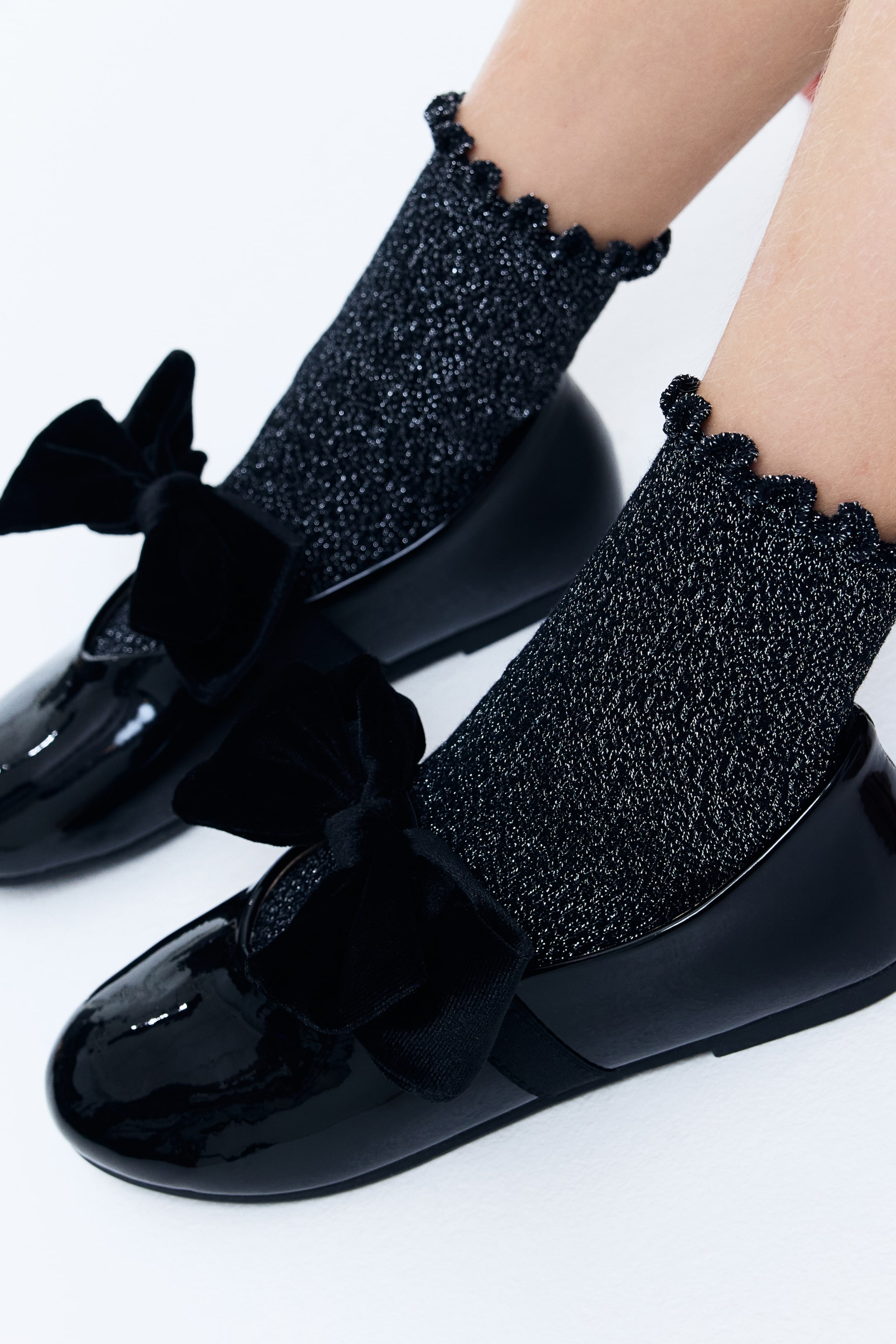 View larger image: A pair of black patent leather flats is being presented from a slightly elevated, front-side angle, featuring a velvet bow on a strap across the vamp. Black glittery ankle socks, with ruffled cuffs, are being worn with the flats, extending above the shoe line.