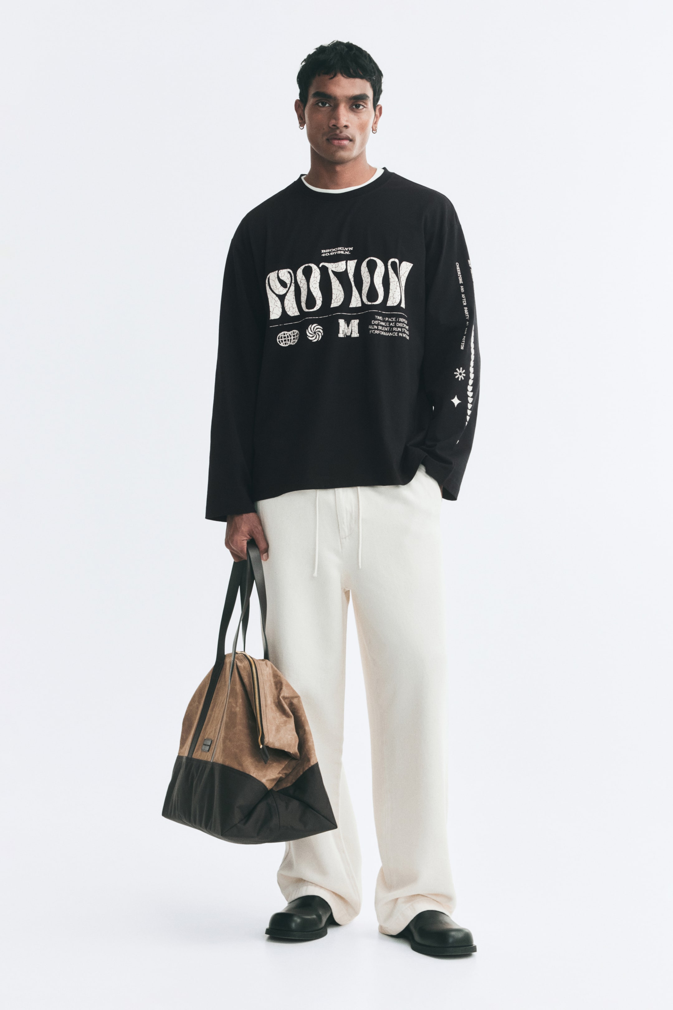 View larger image: A black long-sleeve tee with white printed graphic designs on the chest and sleeve is being worn by the model. The tee features a round neckline, dropped shoulders, and a straight-cut hem, and is paired with cream-coloured trousers.