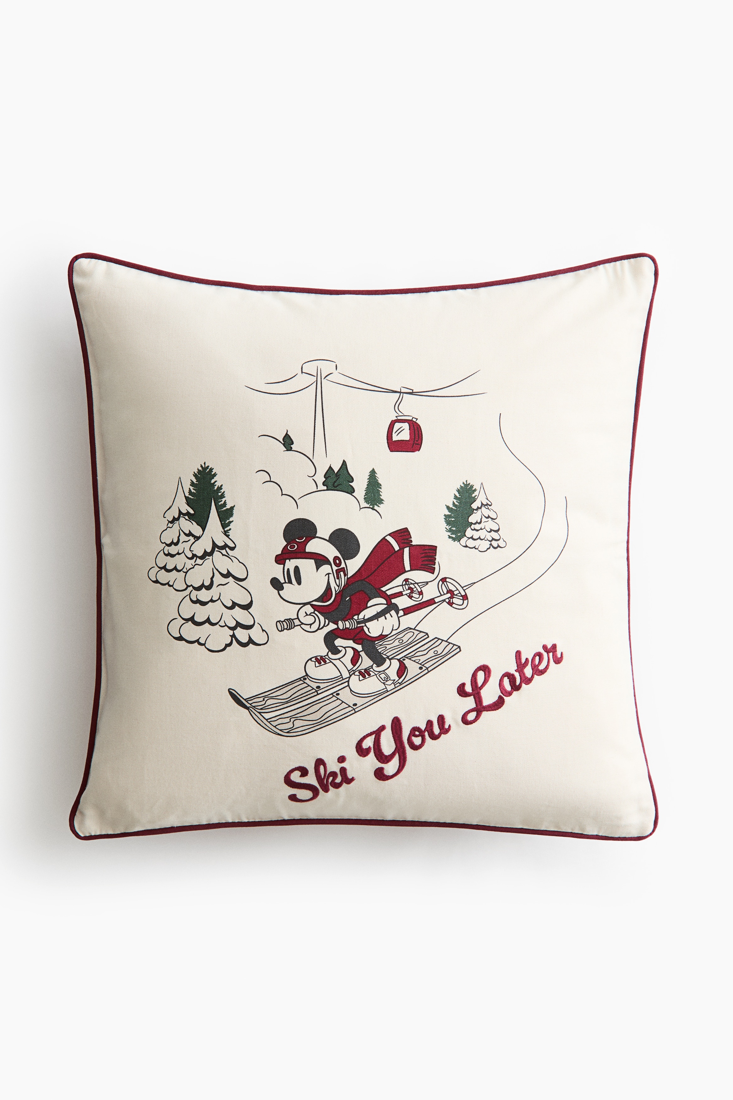 A square cushion cover in off-white cotton canvas is shown from the front, resting upright on a folded beige blanket and a stack of cushions. It features a printed design of Mickey Mouse skiing down a snowy hill next to the embroidered cursive text "Ski You Later" in dark red, with matching dark red piping along the edges.