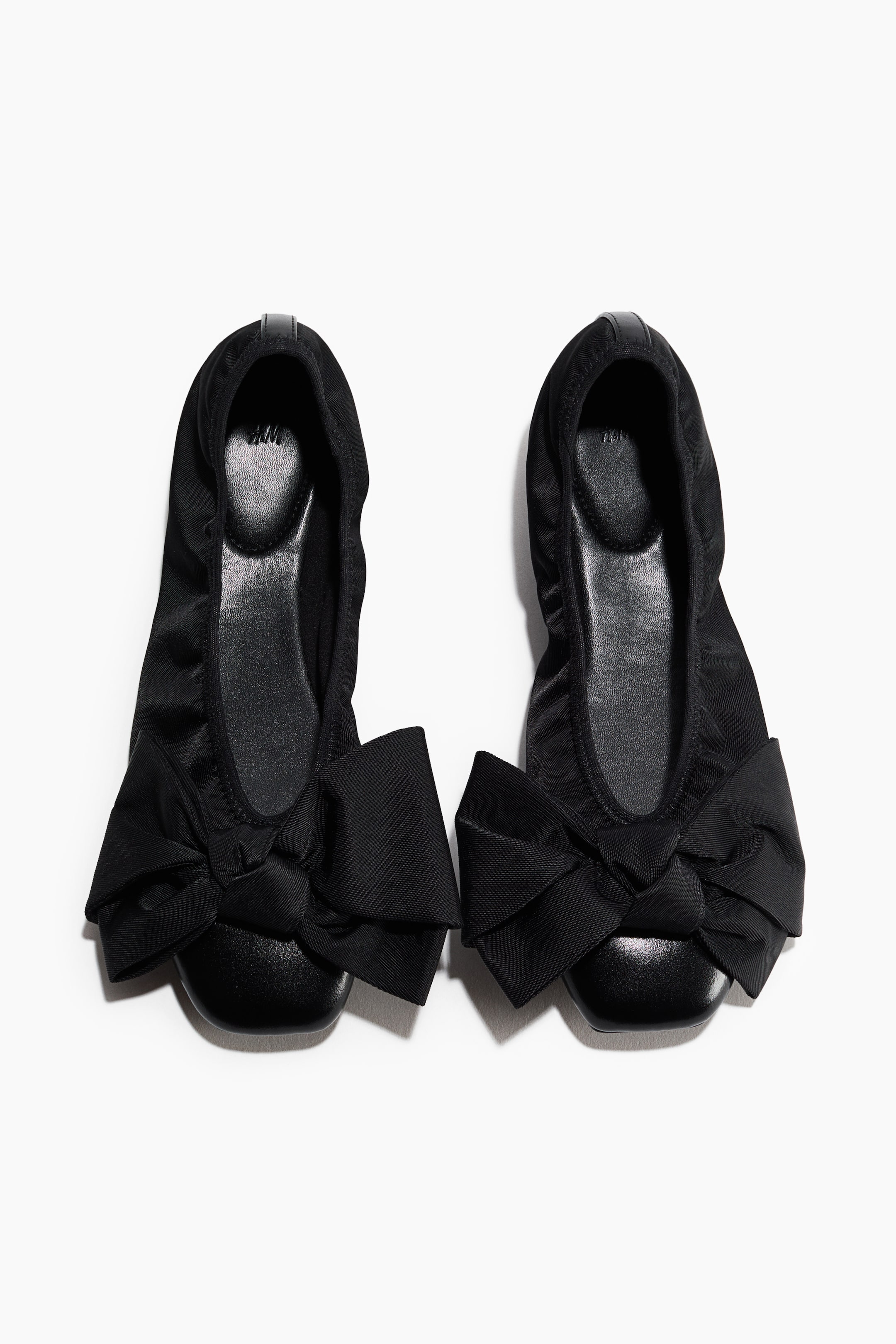 View larger image: A pair of black ballet flats is displayed from a top-down view, featuring coated fabric uppers with elasticated collars and smooth, rounded toe caps. Oversized decorative fabric bows adorn the front of each shoe, which is lined with jersey fabric.