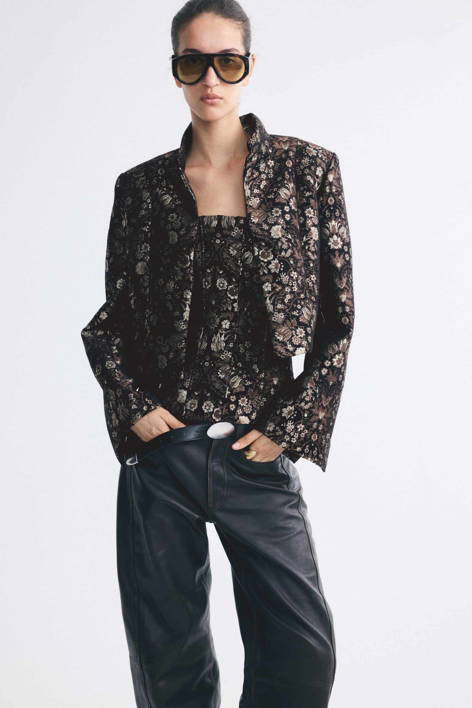 A short jacket in a dark floral brocade weave is worn open by the model, highlighting its structured shoulders and stand collar. The jacket is layered over a matching floral brocade top and styled with dark leather trousers. - 1