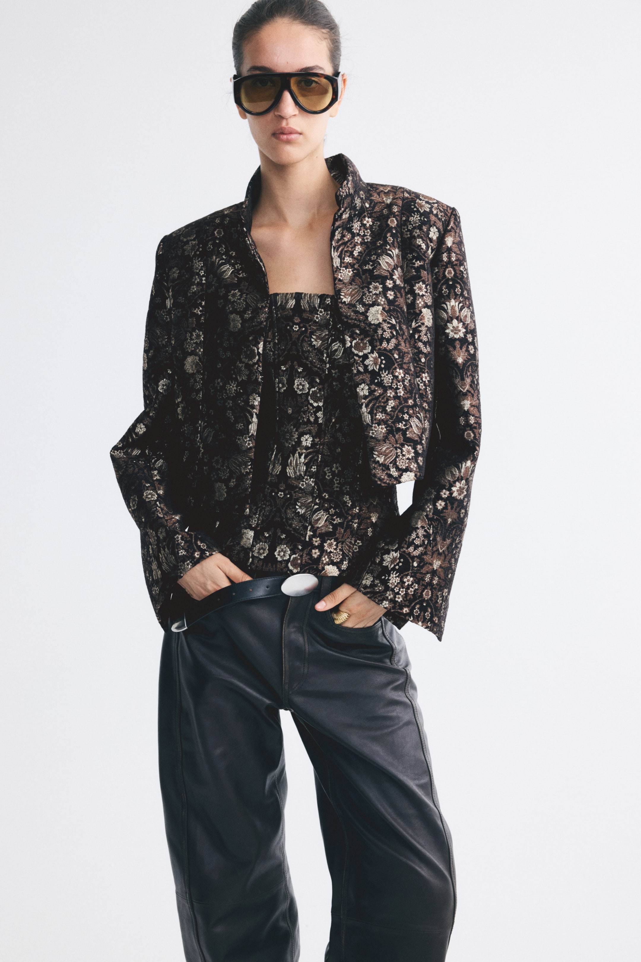 View larger image: A short jacket in a dark floral brocade weave is worn open by the model, showcasing its structured shoulders and stand collar. The jacket is layered over a matching floral brocade top and styled with dark leather trousers.
