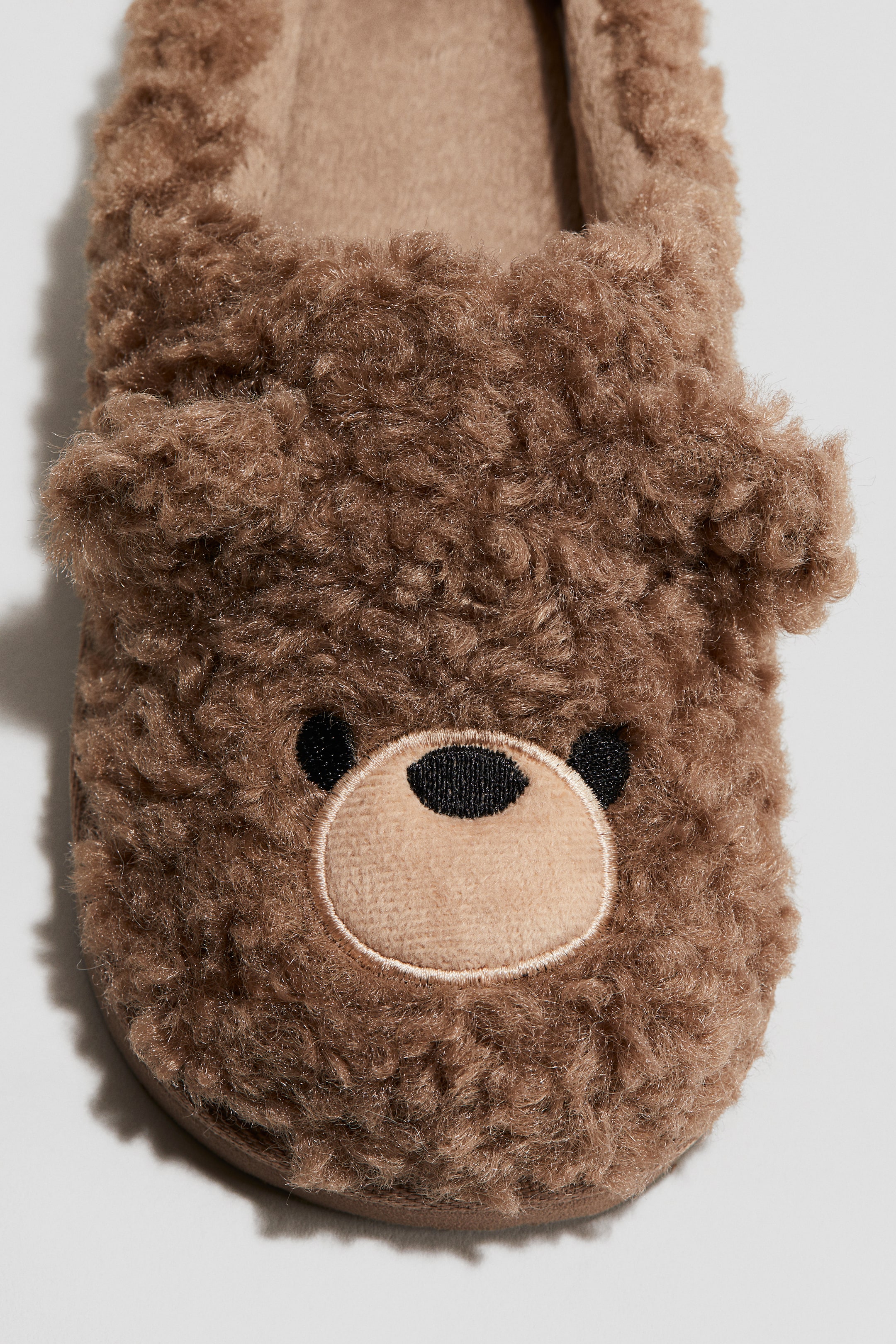 Grotere afbeelding bekijken: Een bruine teddy fleece pantoffel wordt van de VOORKANT bekeken, ontworpen om op het gezicht van een beer te lijken. Het heeft twee kleine ronde oren aan de bovenkant, een lichtere tan ovale snuit met zwarte ronde ogen en een zwarte ovale neus aan de voorkant.