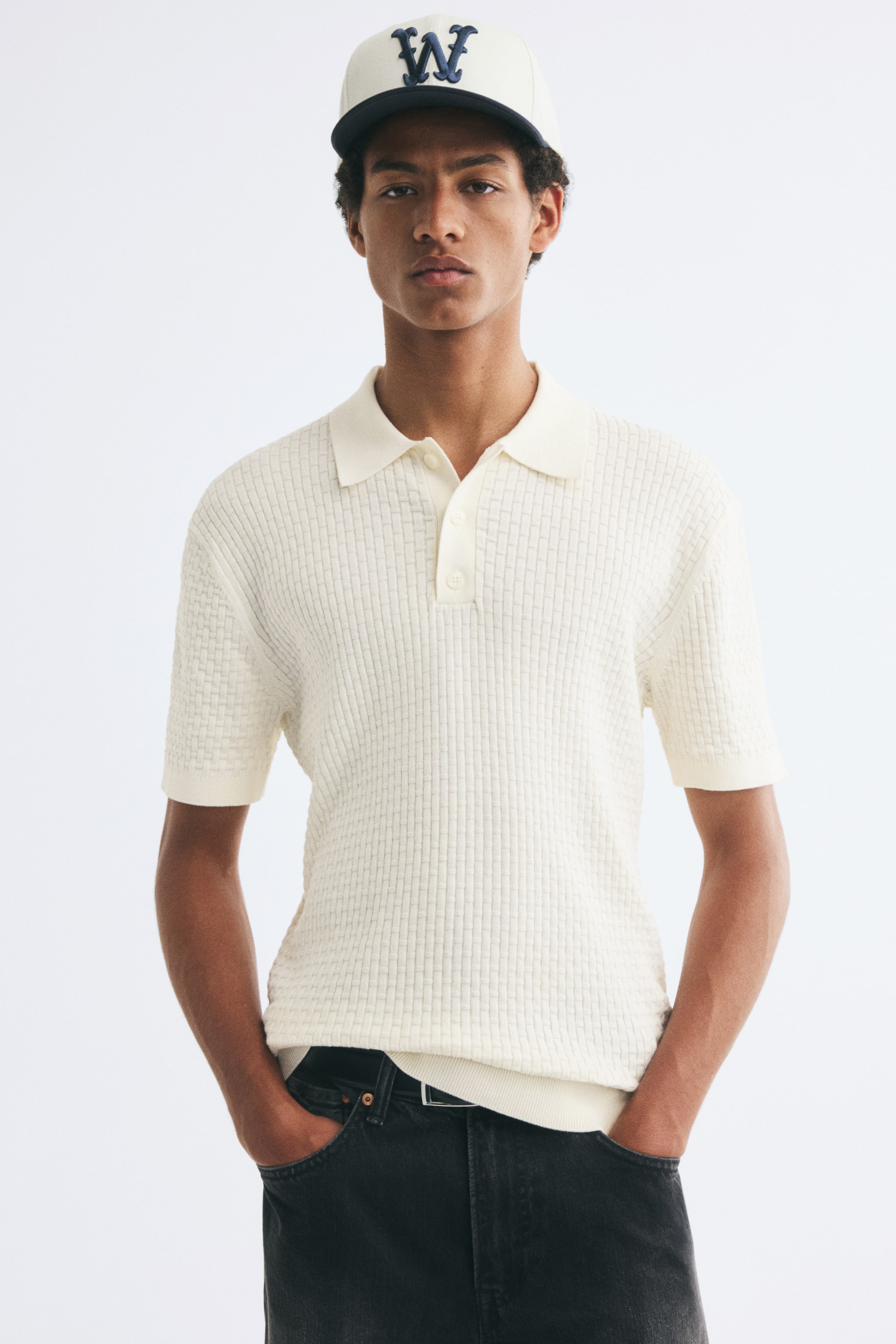 View larger image: A short-sleeve textured knit polo shirt in cream is being worn by the model and styled with dark gray jeans. The shirt features a basketweave-textured pattern, a ribbed collar with a button placket, and fine ribbing at the cuffs and hem.